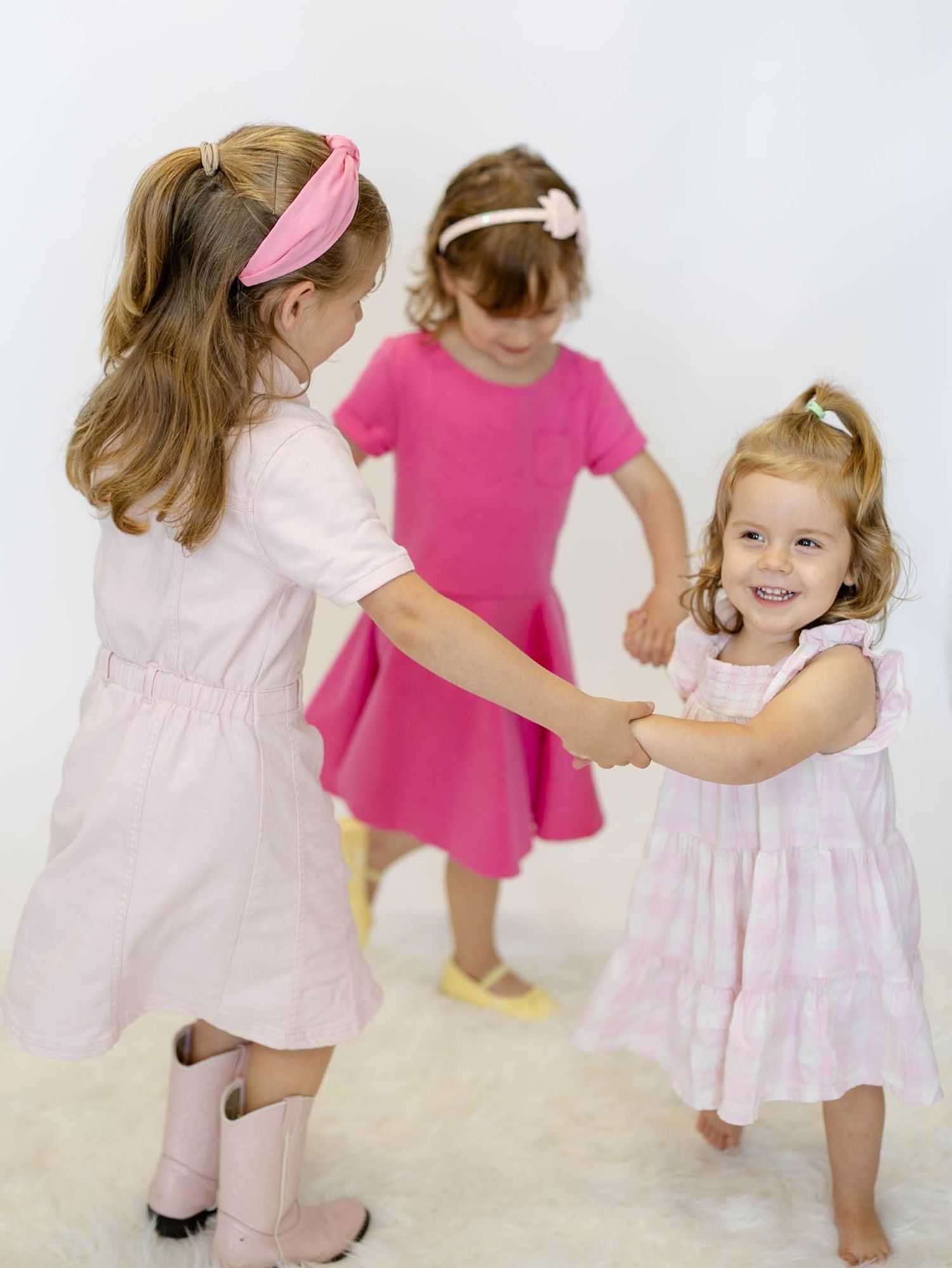 Three young girls in pink dresses dancing and holding hands together in a bright white studio setting.
