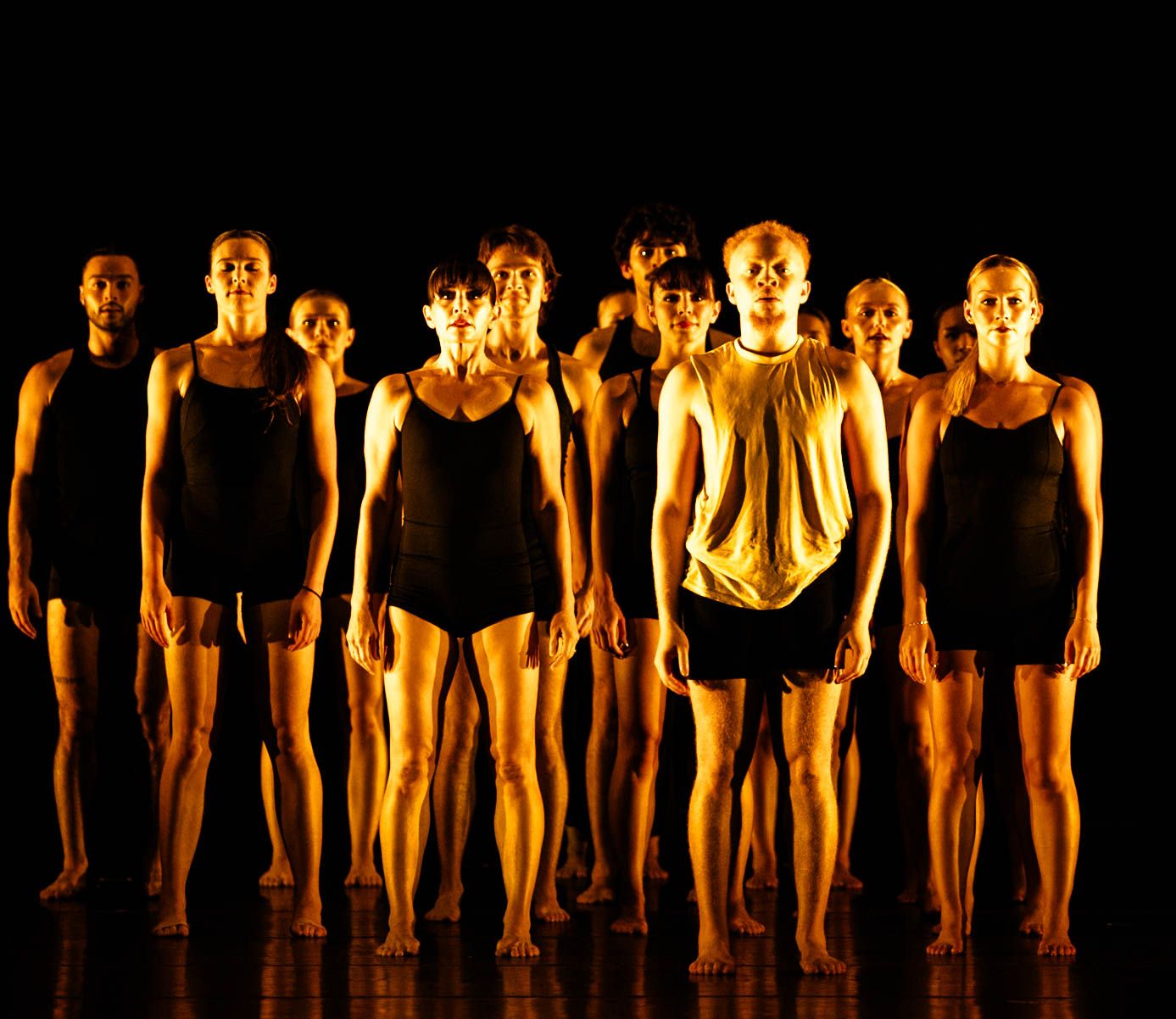 A group of barefoot contemporary dancers in dark athletic costumes stand close together, lit in warm golden light against a black stage.
