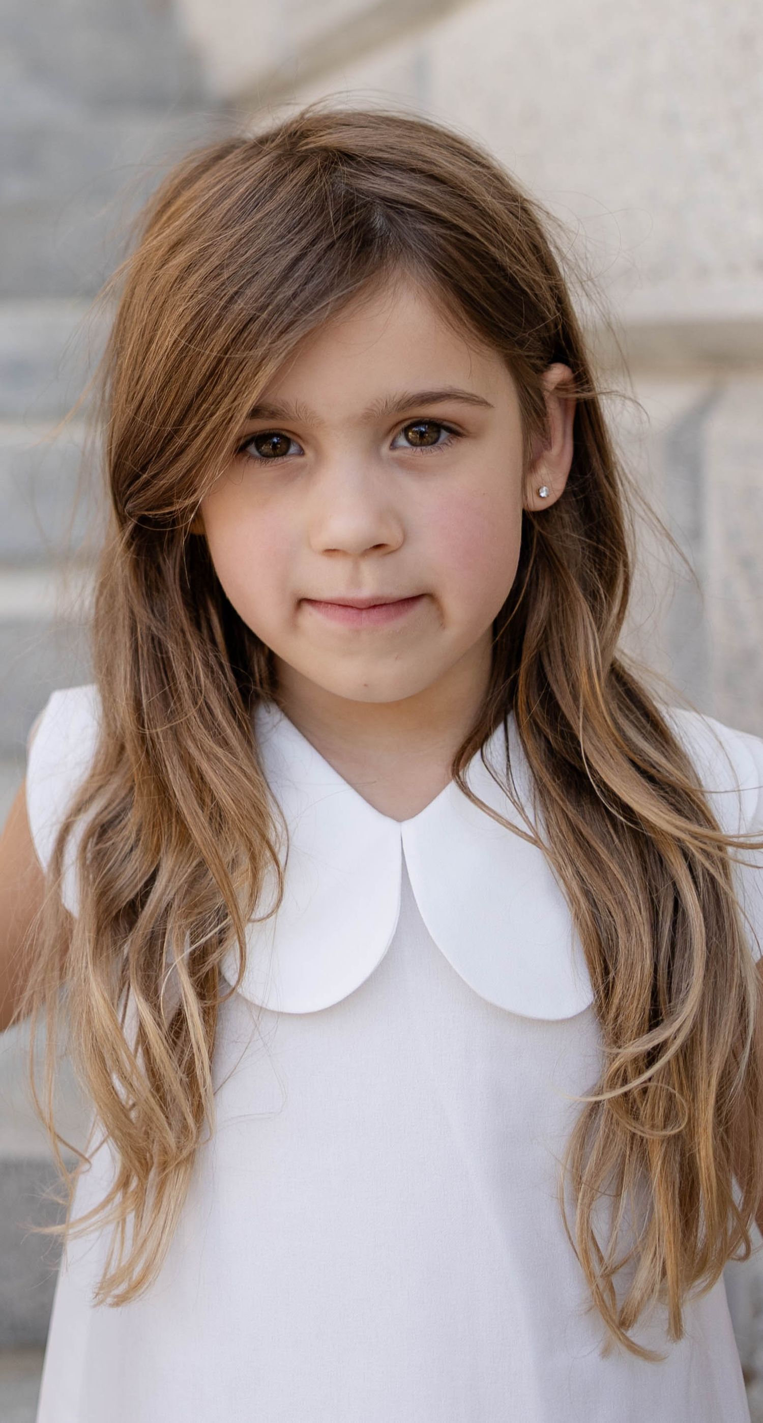 A pretty young girl with long brown hair wearing a white Peter Pan collar dress poses for a portrait.