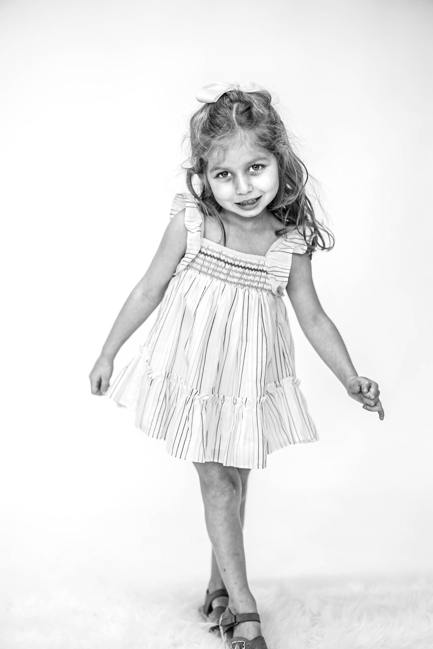 Black and white portrait of a young girl in a striped dress, smiling and posing playfully against a white background.