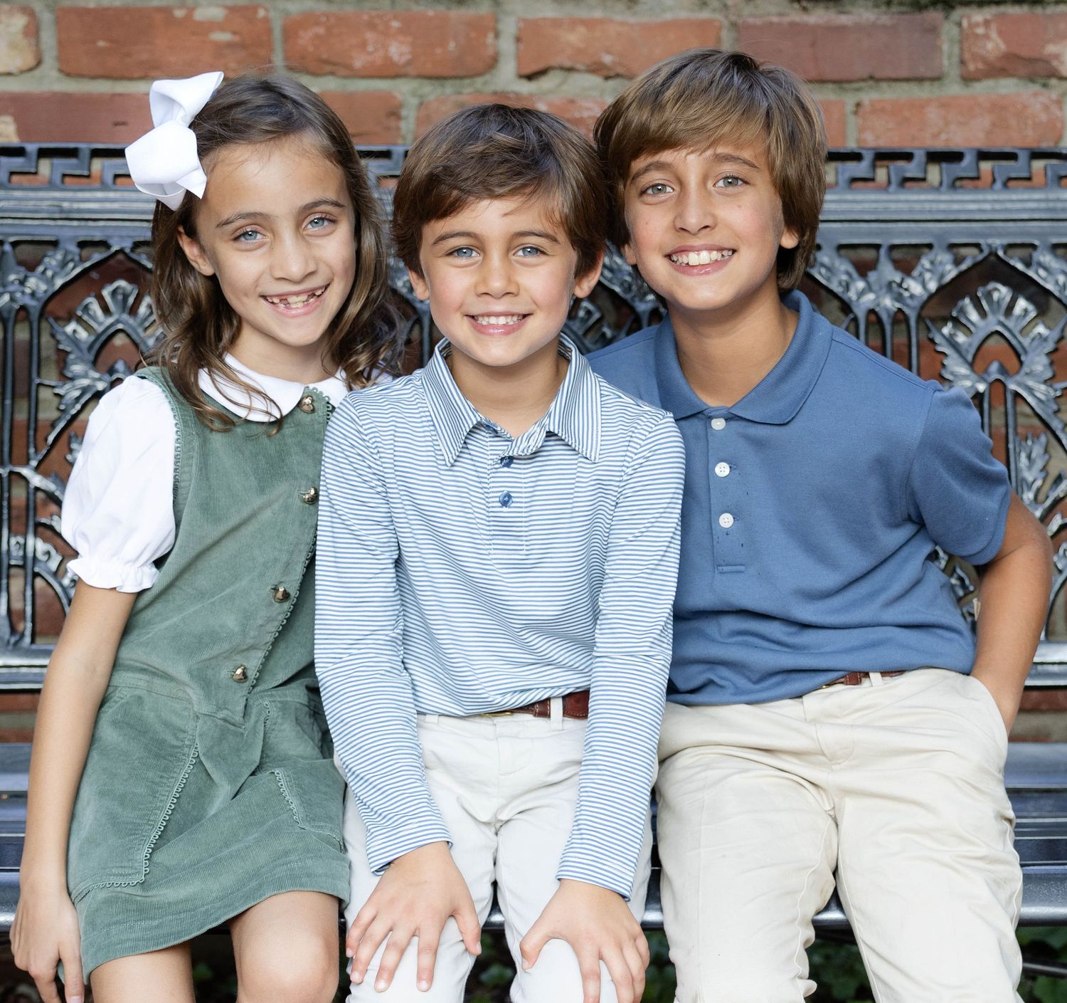 Three smiling children sit together on an ornate metal bench in front of a brick wall, a girl in a green dress and two boys in polo shirts.
