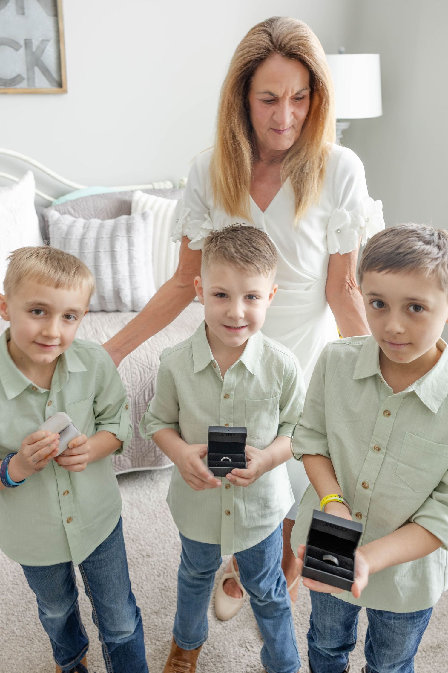 A mother in white dress stands behind three boys in matching green shirts holding ring boxes in a bright bedroom.