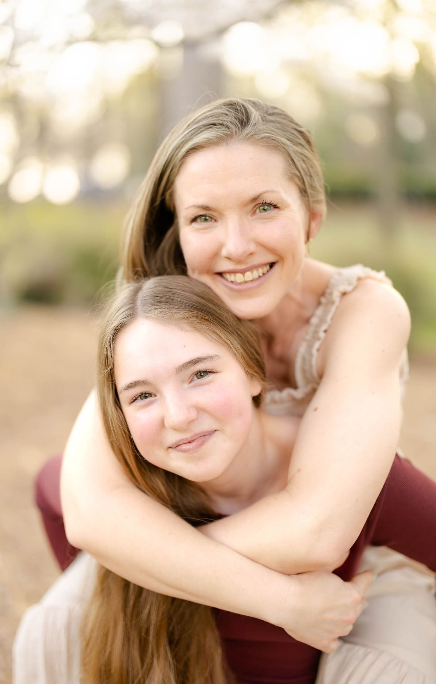 A smiling woman embraces a teenage girl from behind in a bright, bokeh-filled outdoor portrait.