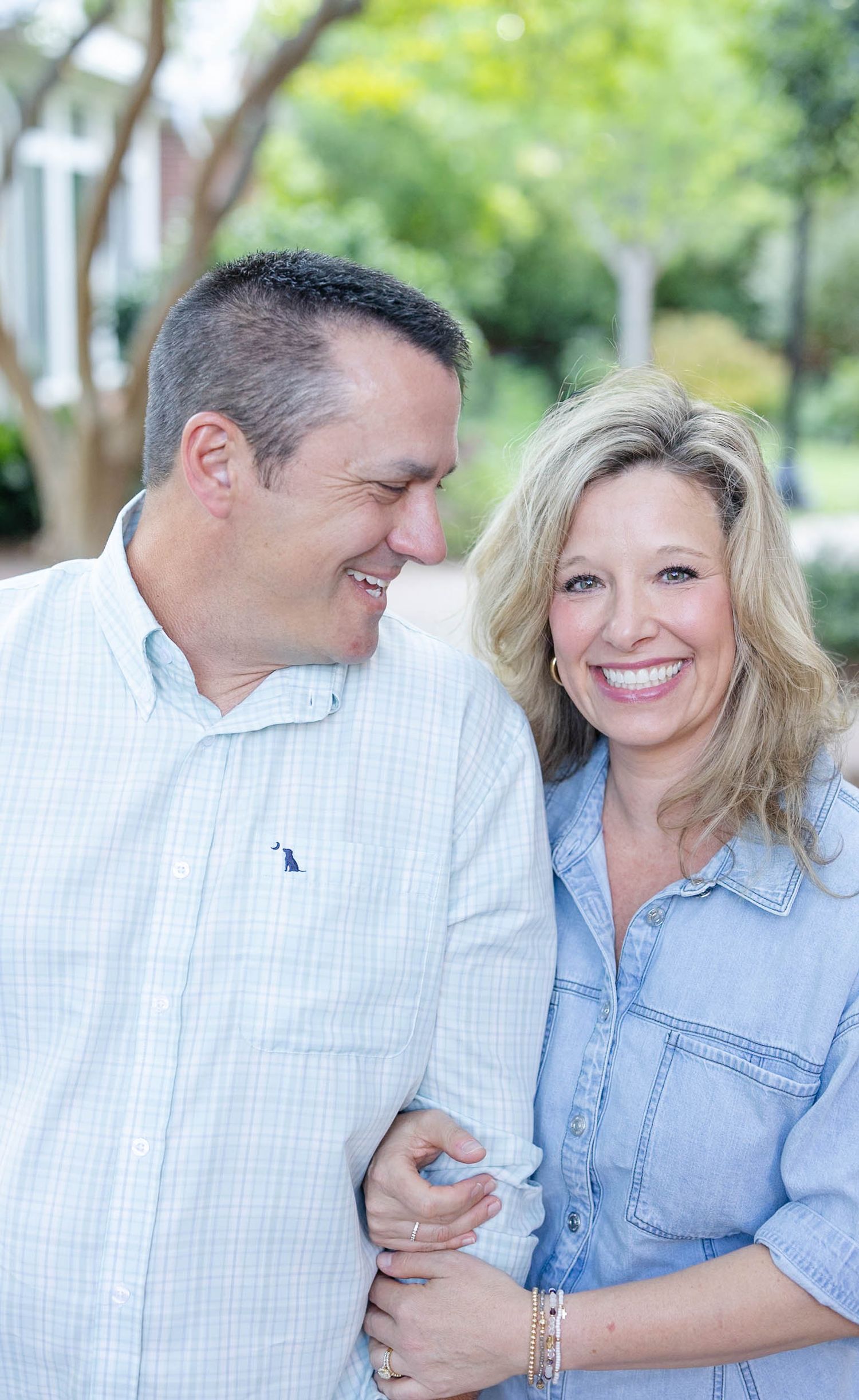 Smiling couple embracing outdoors, woman in denim shirt and man in light blue plaid shirt, posing among green trees.