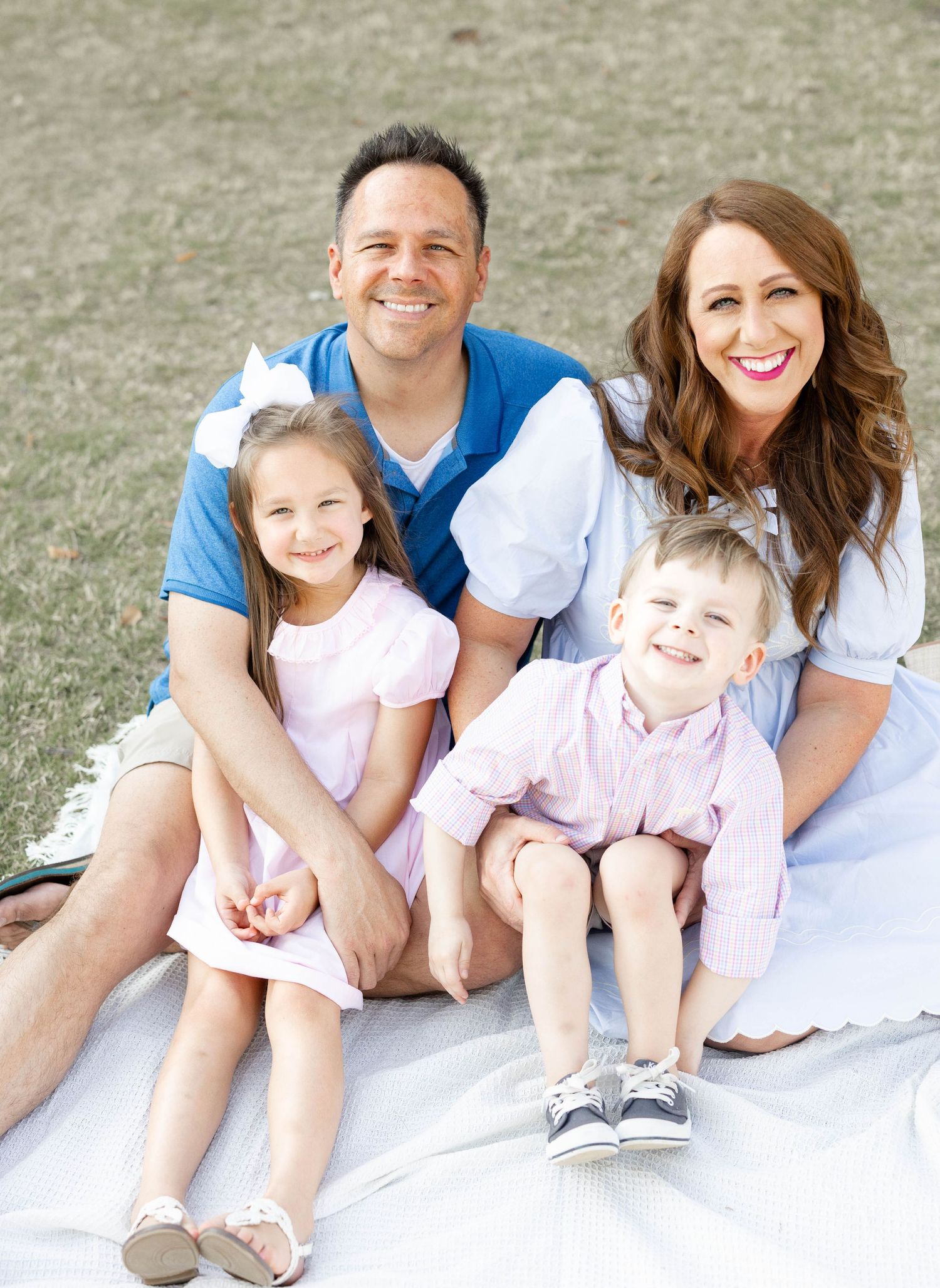 Joyful family of four sits on blanket outdoors, parents and two young children smiling.