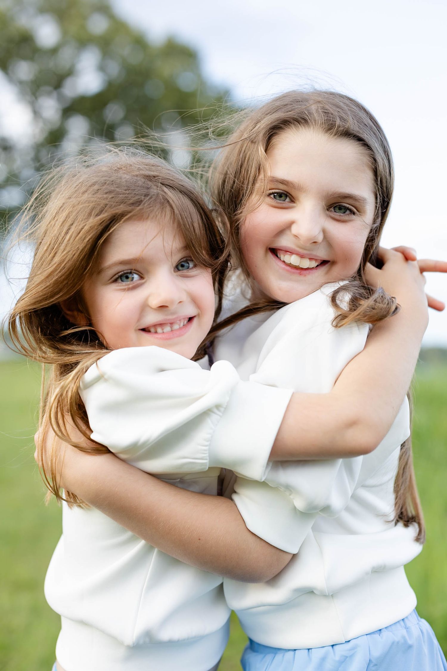Two young girls with brown hair hugging and smiling outdoors in white tops.