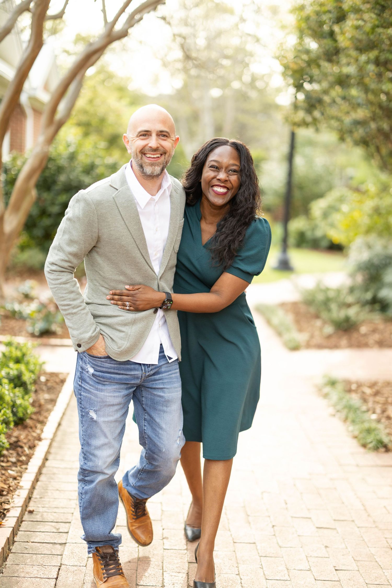 Smiling interracial couple embracing on a sunlit brick pathway surrounded by green trees.