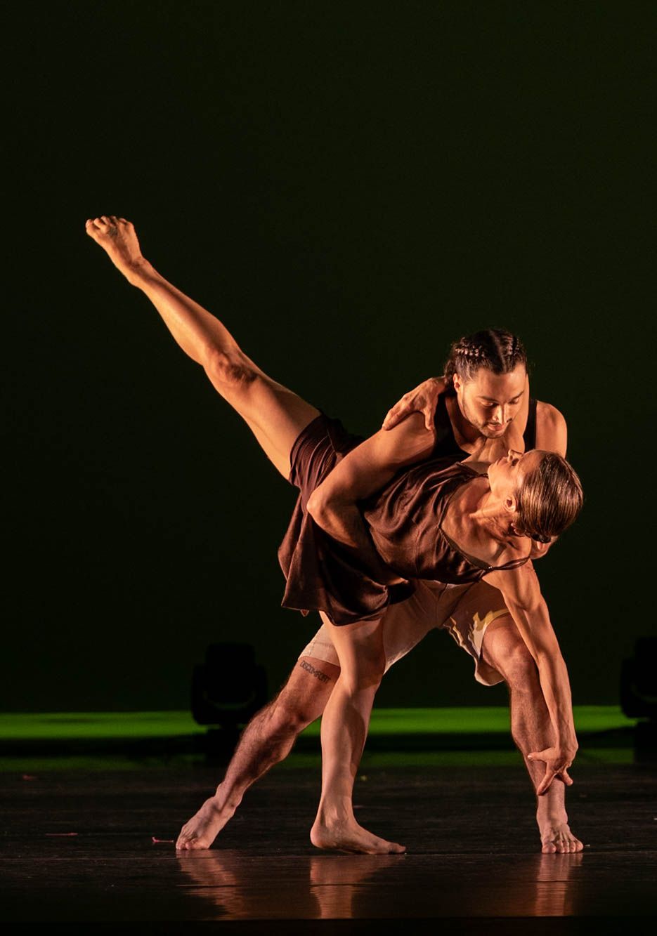 Two barefoot contemporary dancers perform a dramatic partnered lift on a dark stage, one leaning over the other with a leg extended high.