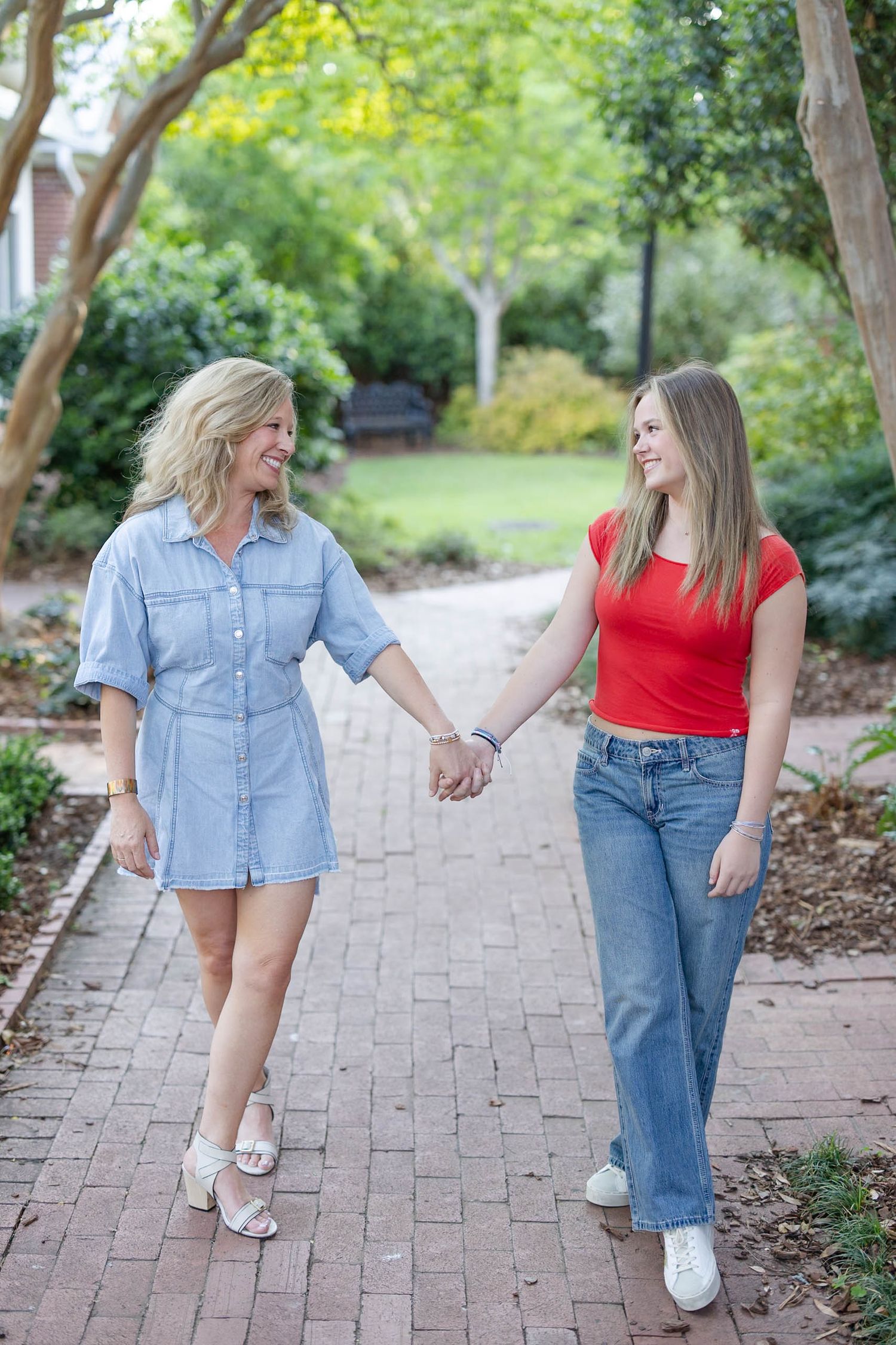 Blonde woman in denim dress and teen girl in red top holding hands on a brick garden pathway.