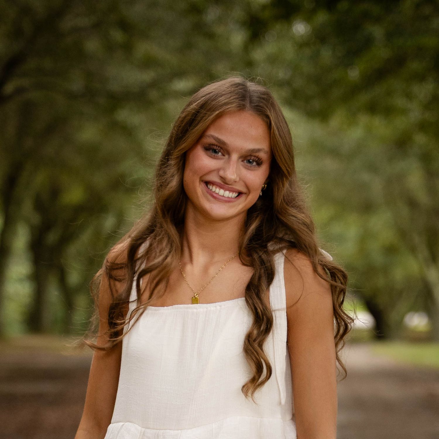 Smiling young woman with long wavy brown hair wearing a white sleeveless top stands on a tree-lined path with soft green foliage behind her.