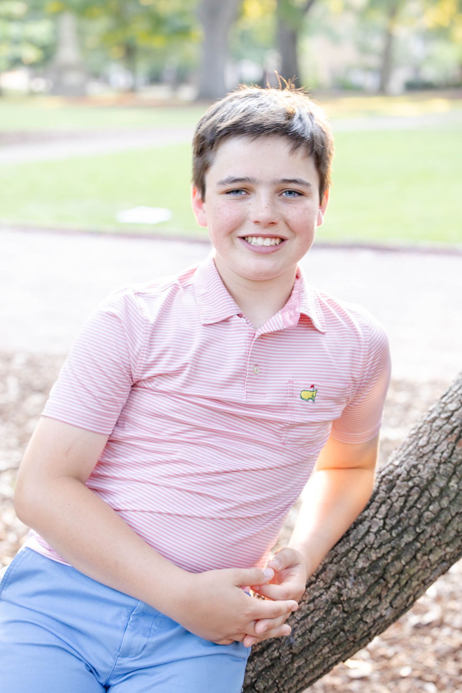 Smiling young boy in pink striped polo shirt leaning against a tree in a sunny park setting.
