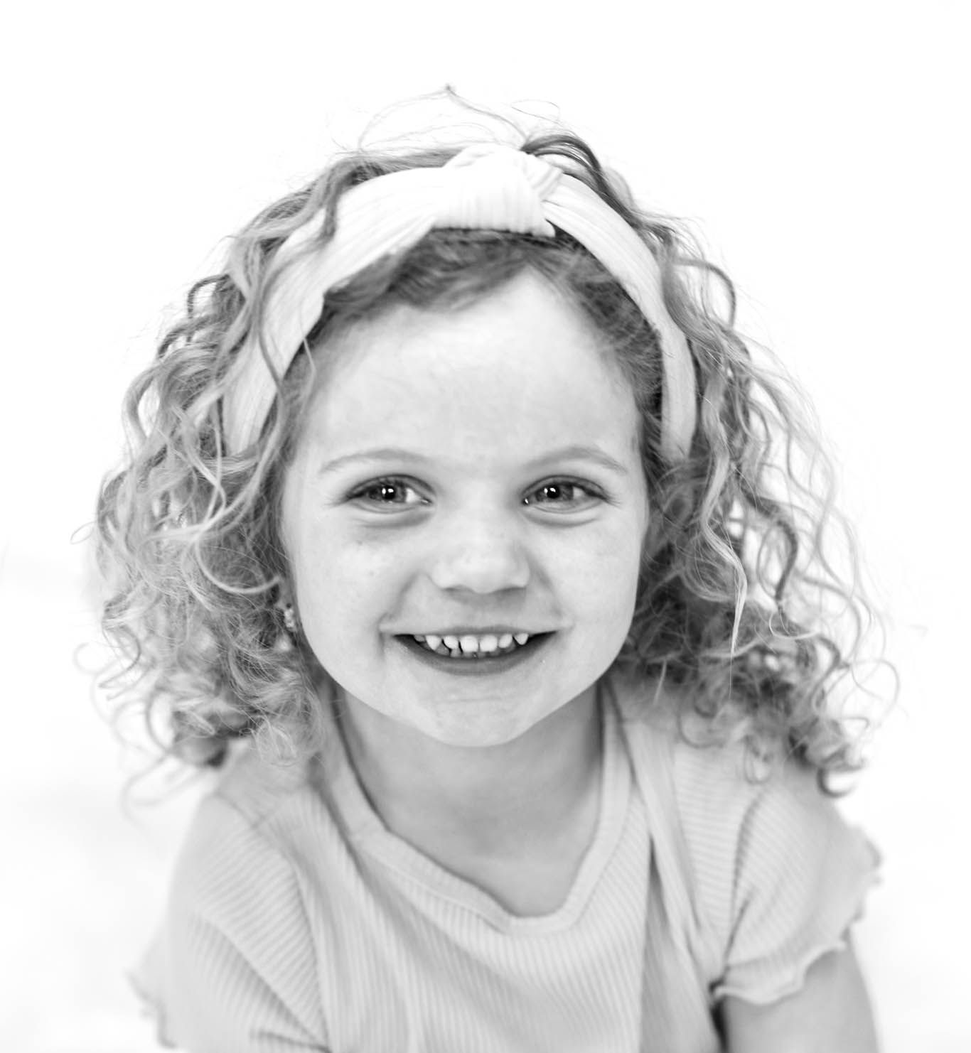 Black and white portrait of a smiling young girl with curly hair and a bow headband.