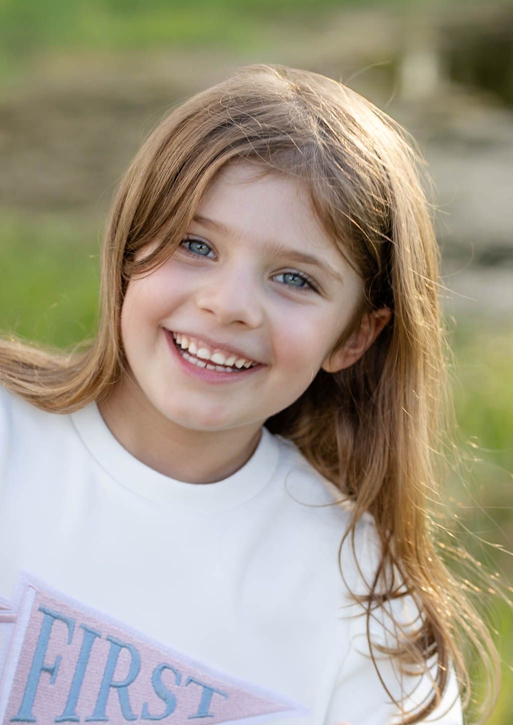 Smiling young girl with long brown hair and blue eyes wearing a white shirt outdoors in natural light.