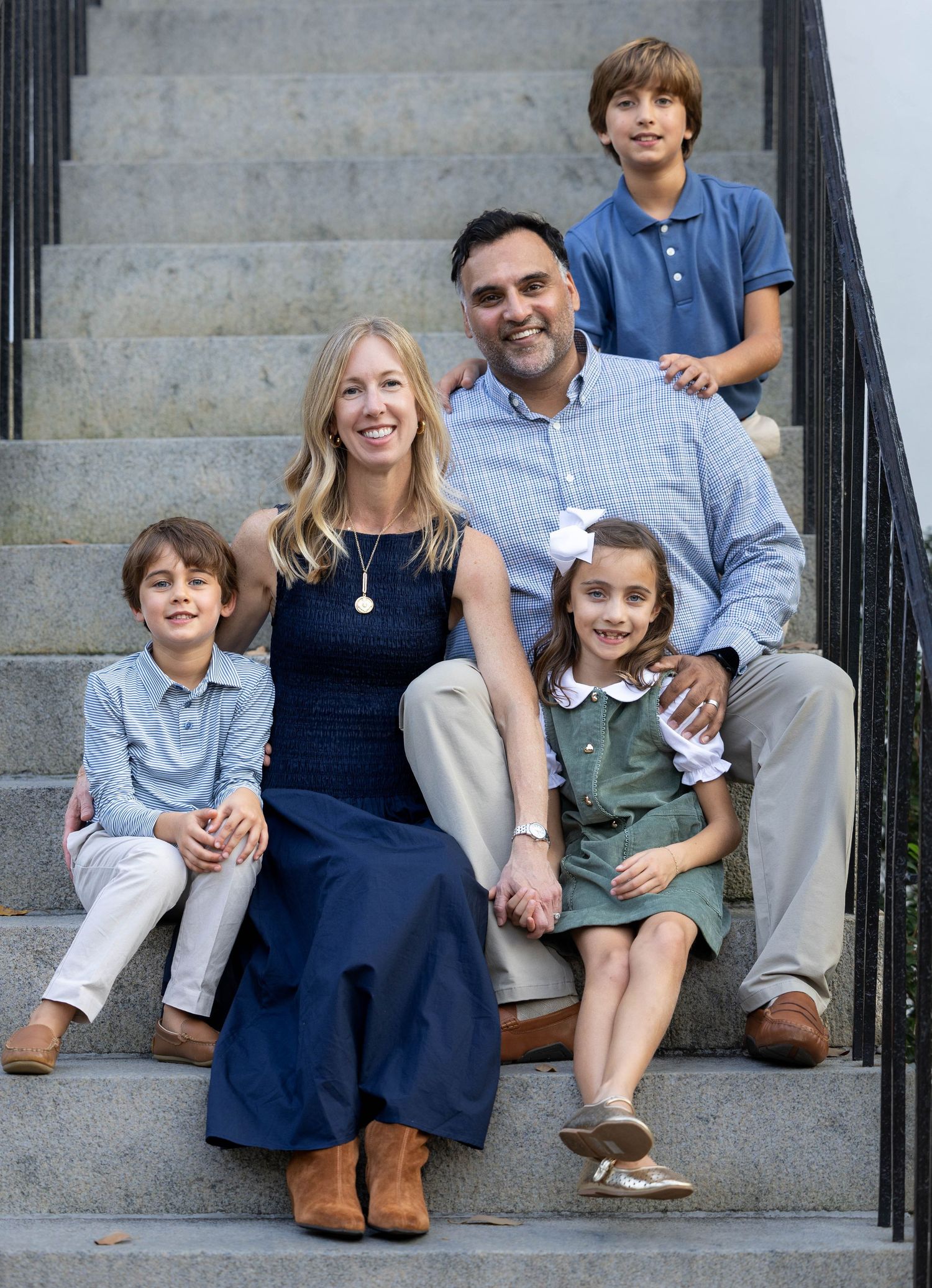 Smiling family of five posing together on outdoor stone steps, with two parents and three children in coordinated blue and neutral outfits.