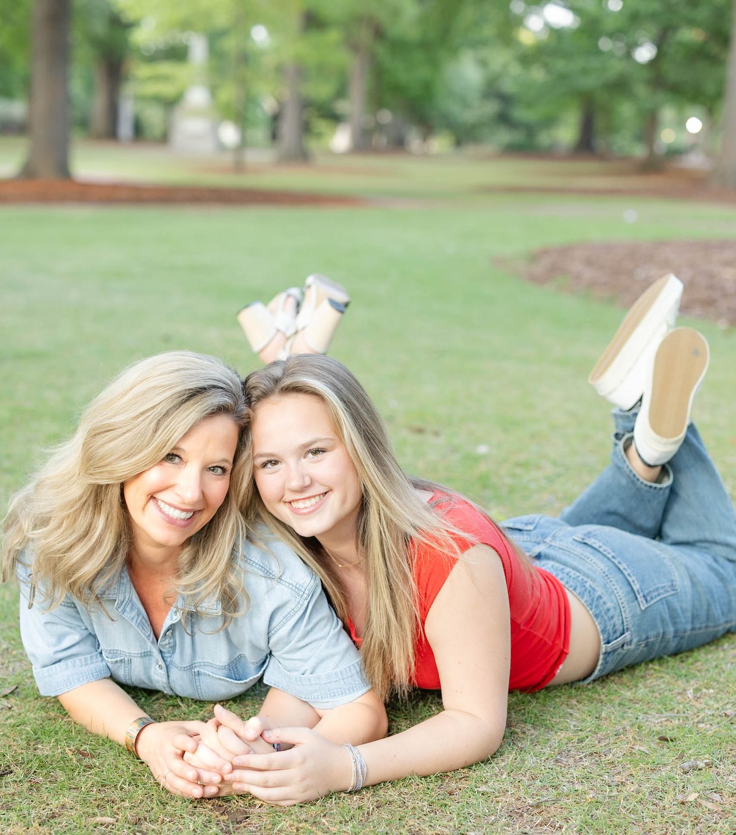 Mother and teenage daughter lying on green grass smiling together in a sunny park setting.