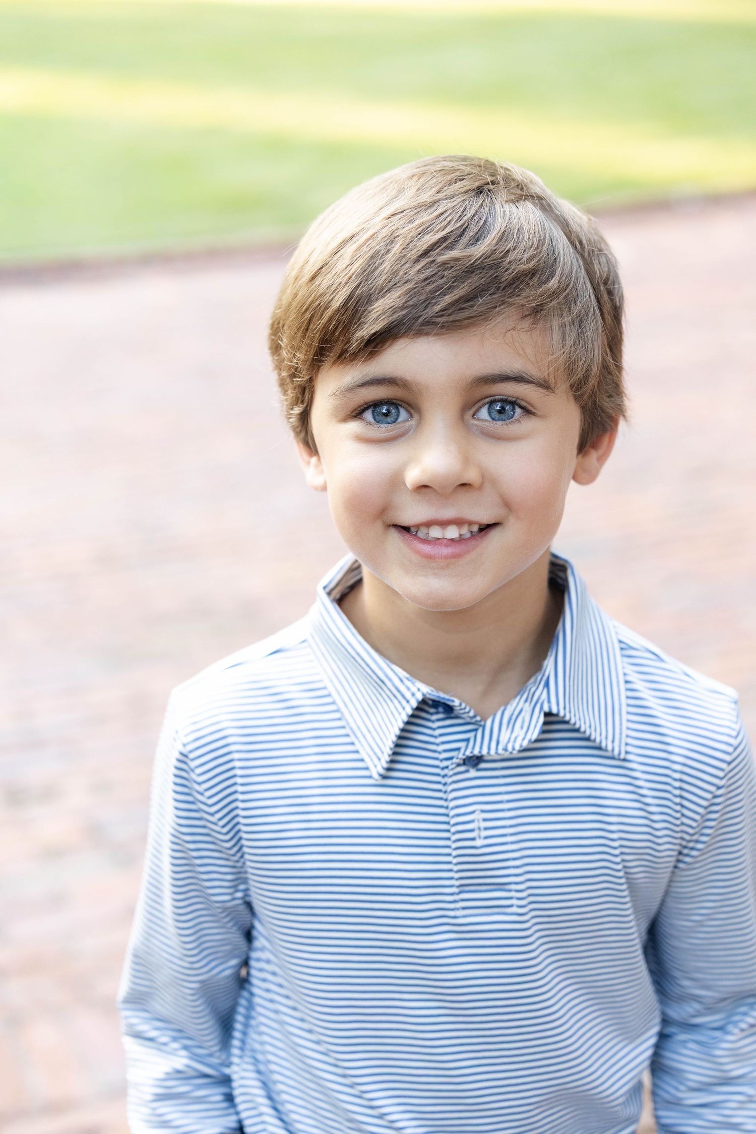 Smiling young boy with light brown hair and blue eyes wearing a blue and white striped collared shirt, standing outdoors on a brick path.