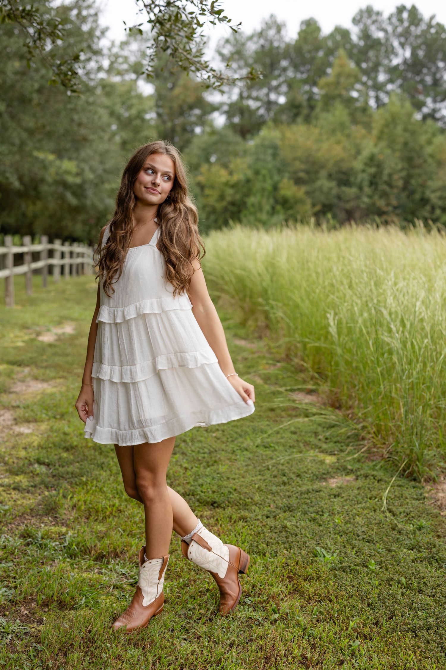 Young woman with long wavy hair in a white tiered sundress and cream cowboy boots stands smiling on a grassy path beside tall field grass.