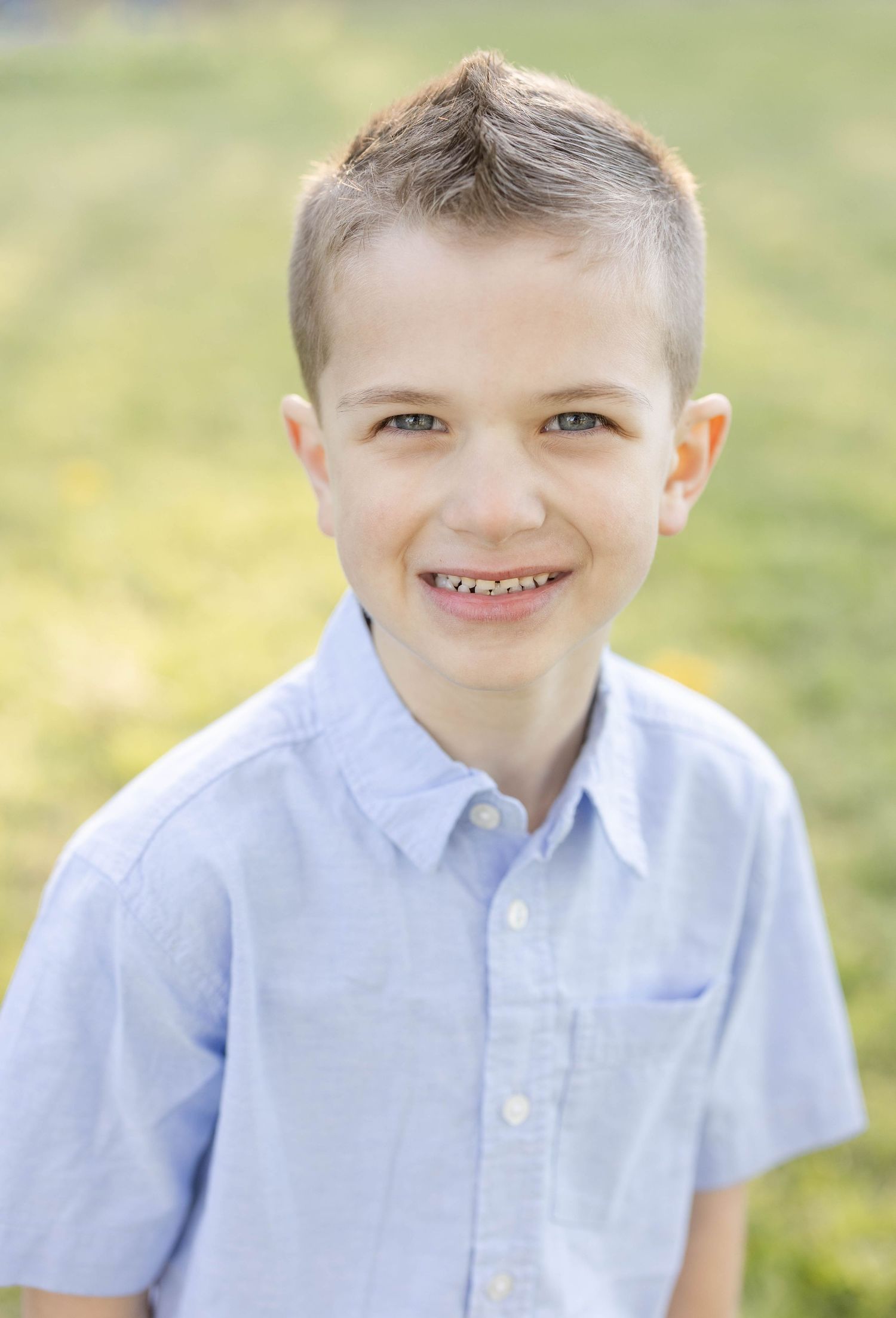 A cheerful young boy with short hair smiling brightly in a light blue button-up shirt outdoors.