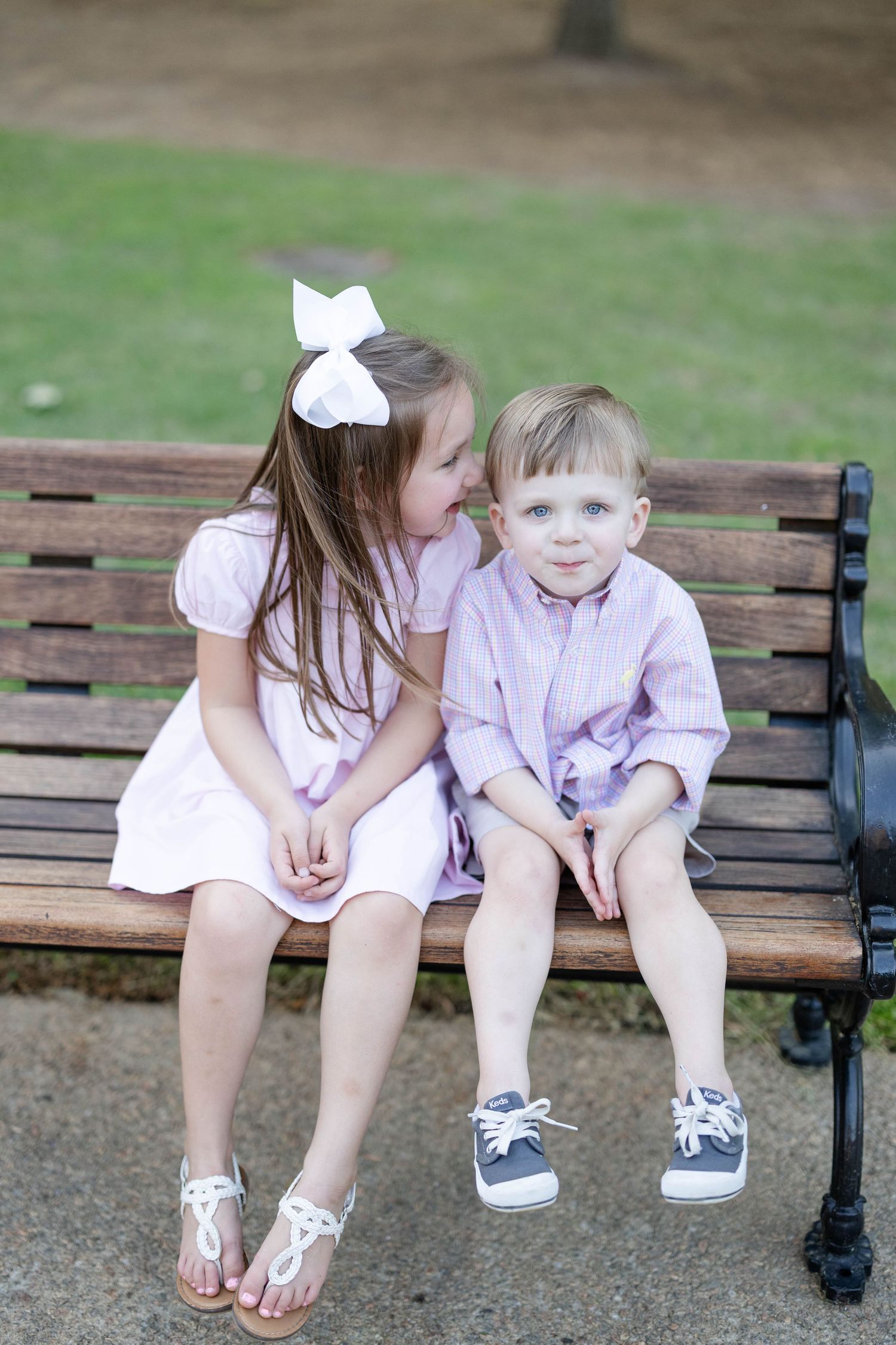 Adorable young girl in pink dress whispers to little boy on a wooden park bench outdoors.