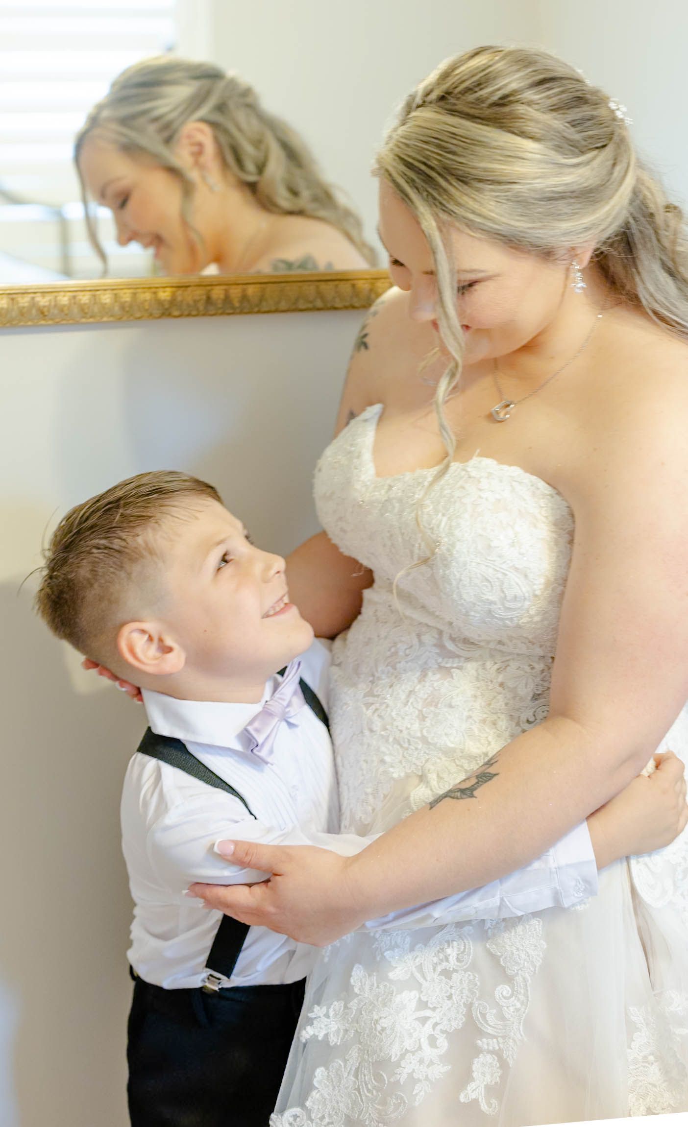 A bride in a white lace gown shares a sweet moment with a young boy in suspenders near a gold-framed mirror.