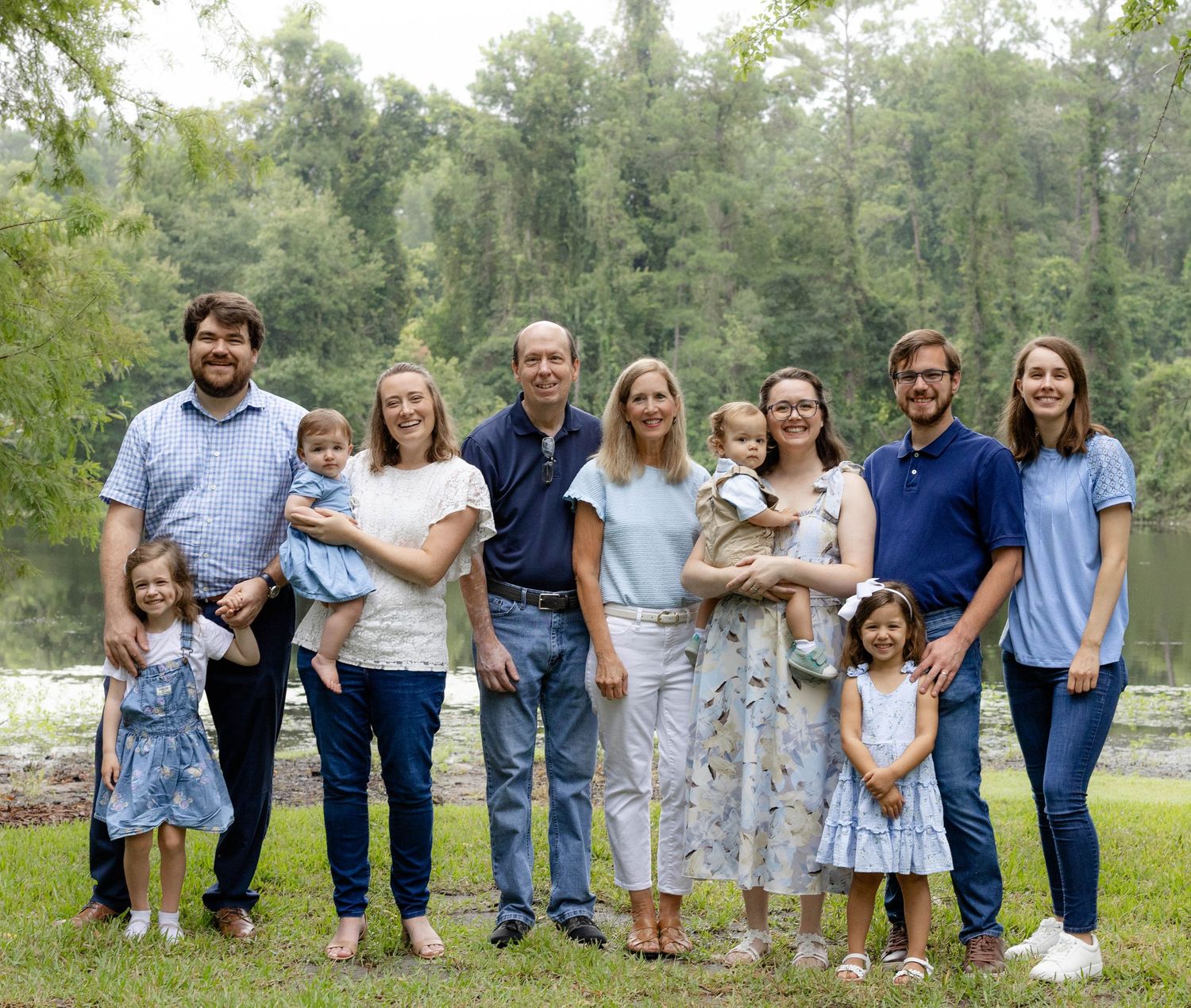 Large family of adults, young children, and toddlers in matching blue and white outfits posing together on a grassy lawn by a river.