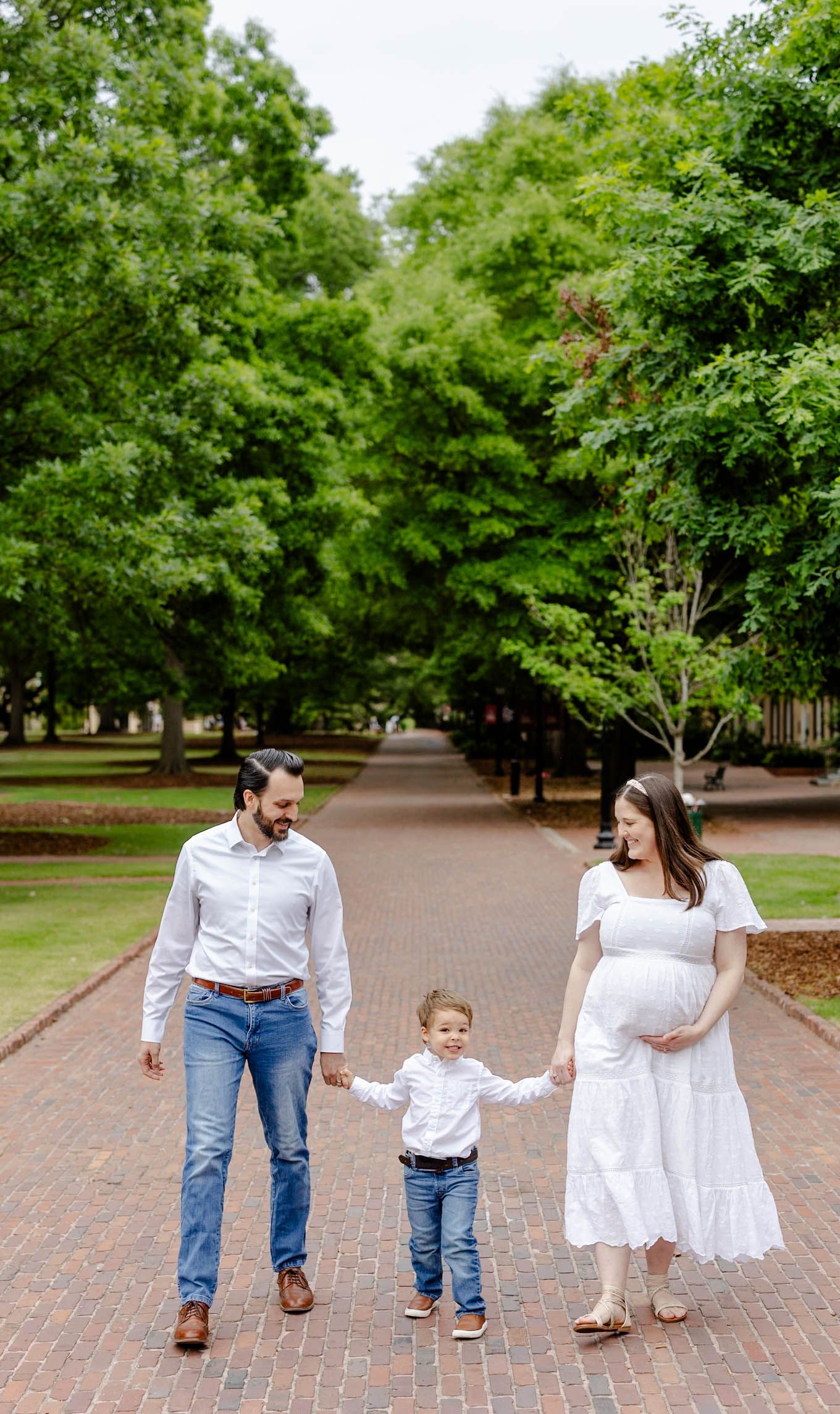Pregnant woman in white dress walks with husband and toddler son on brick path surrounded by green trees.