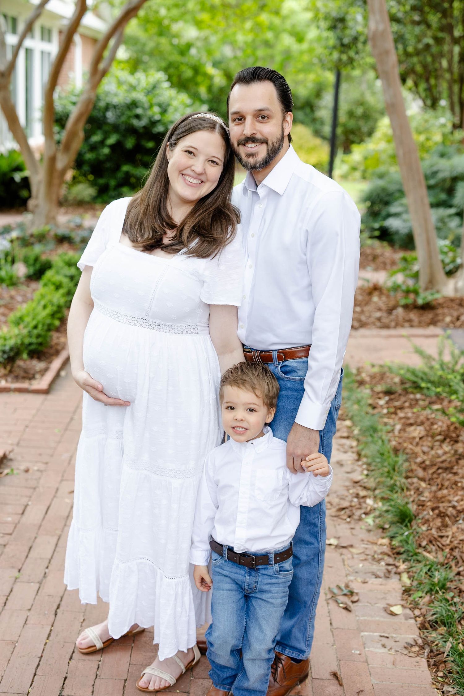 Pregnant woman in white dress poses with husband and young son on brick path in lush garden setting.