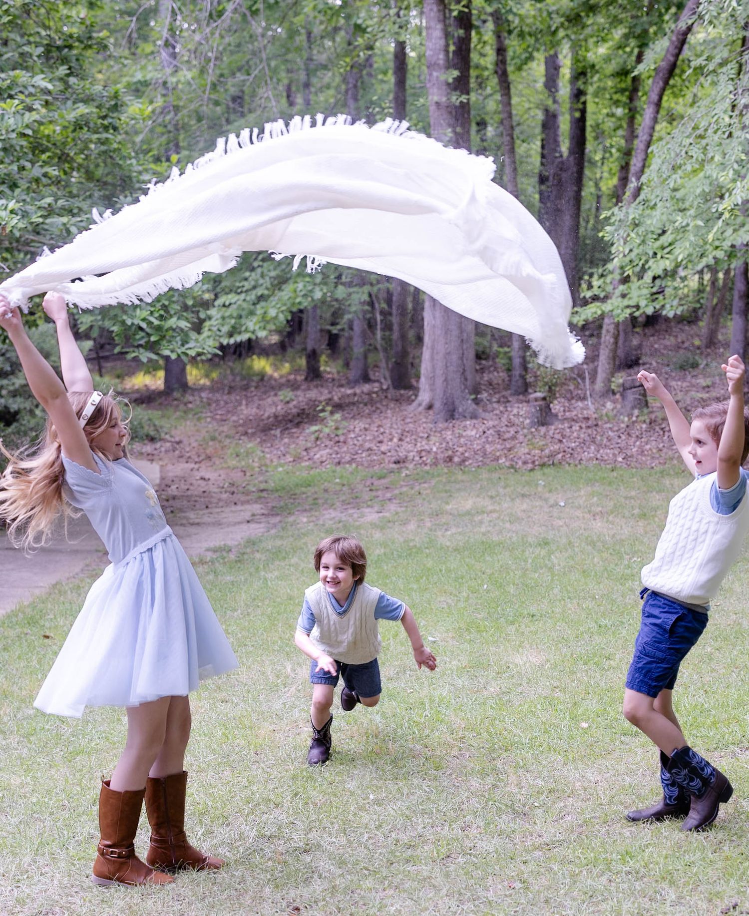 Children playing outdoors with a white blanket flying in the air on a grassy lawn surrounded by trees.