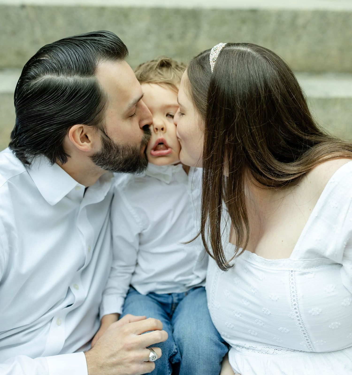 A happy family with two parents in white shirts kissing their young toddler child on the cheeks outdoors.