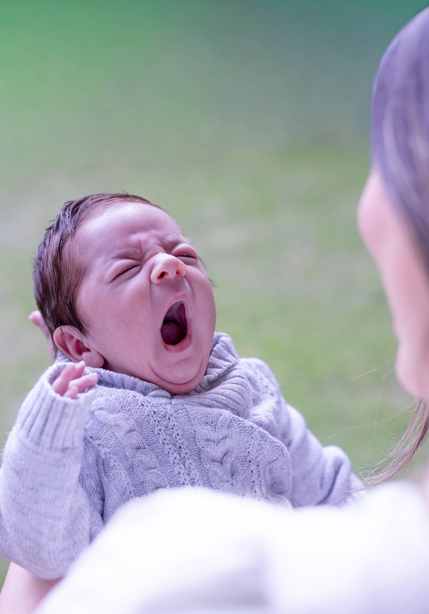 Newborn baby yawning while being held by mother outdoors, wearing a gray knit sweater.