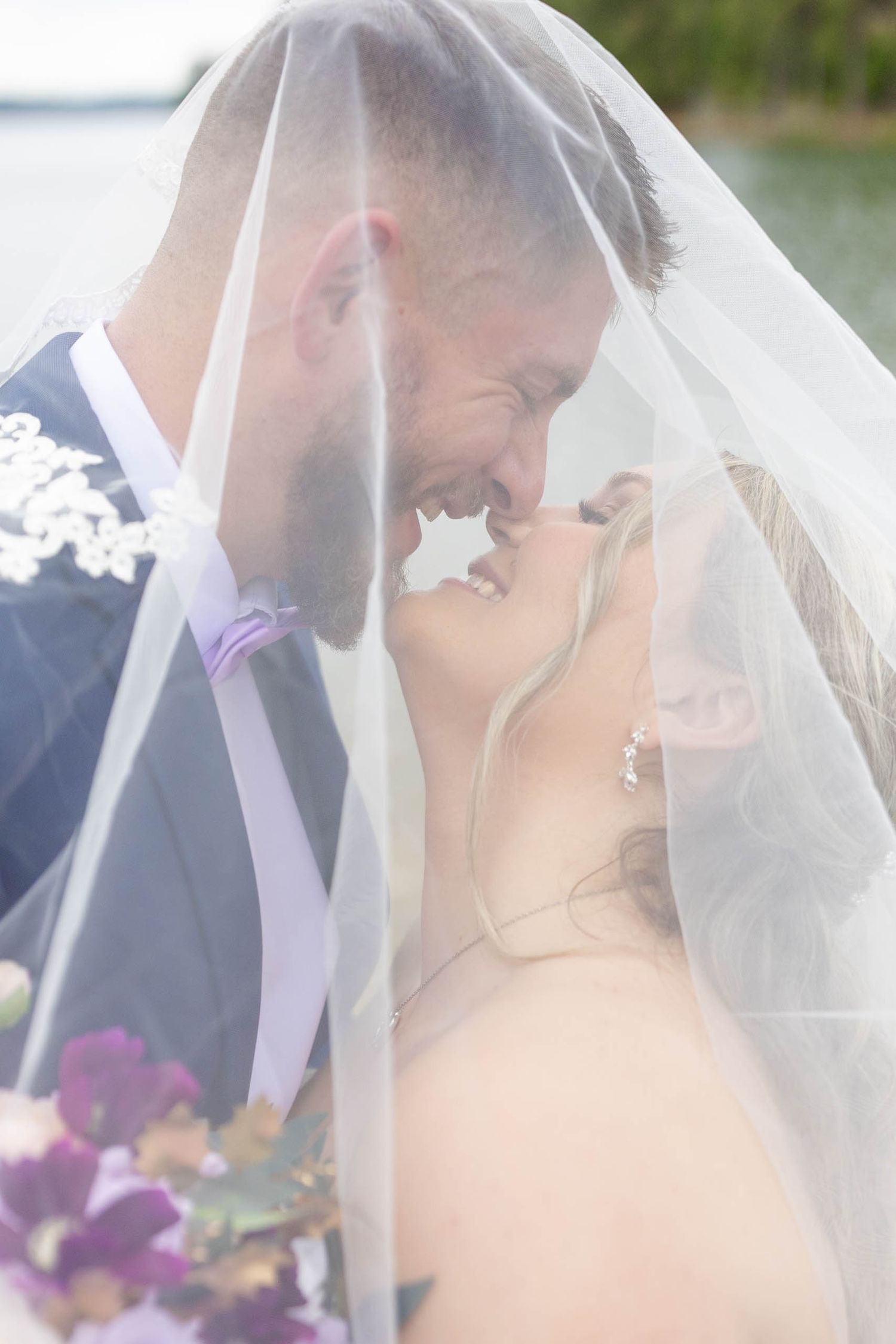 Bride and groom sharing a romantic moment under a wedding veil near waterfront, with purple bouquet.