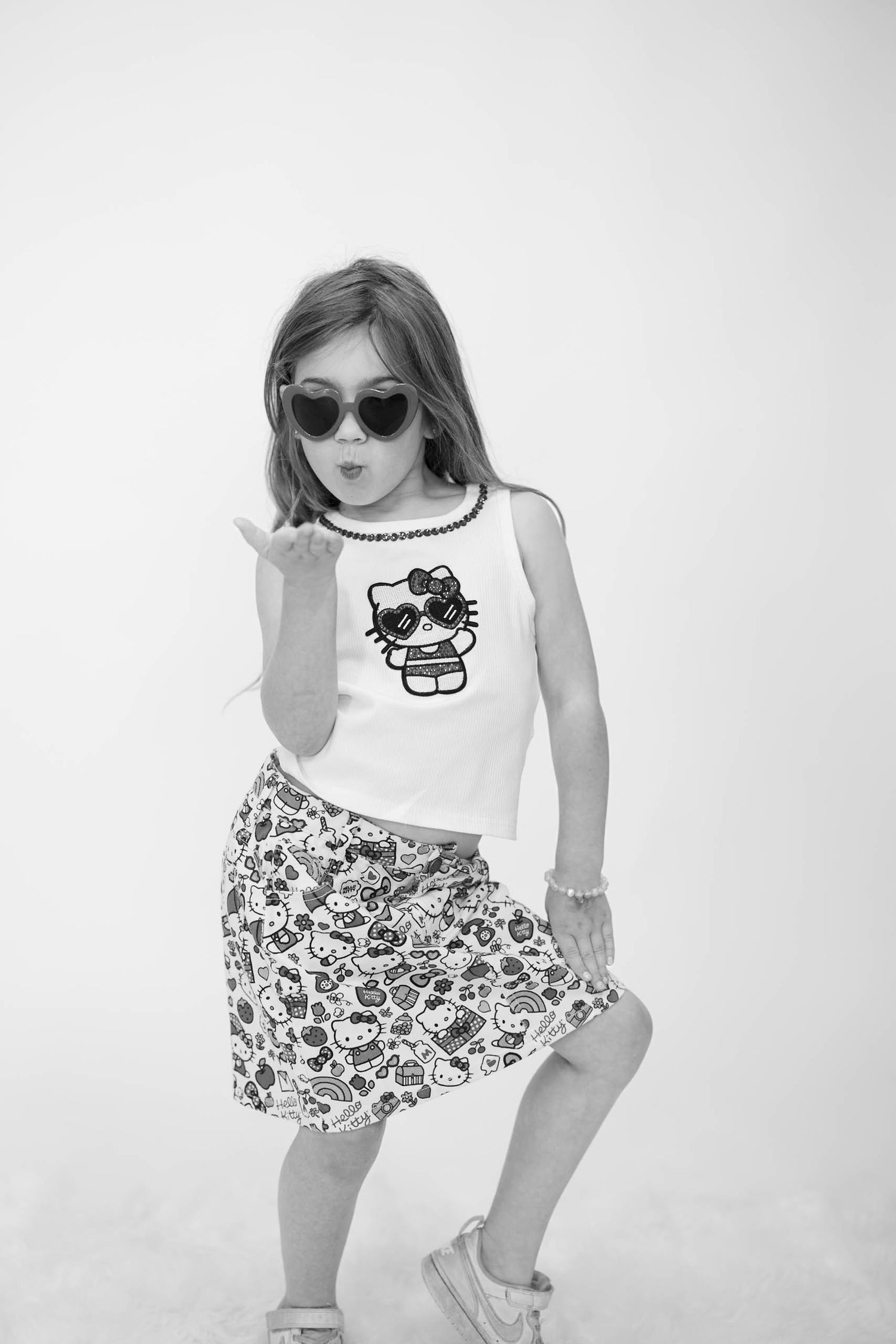 Young girl in Hello Kitty tank top and skirt, heart sunglasses, blowing a kiss in black and white photo.