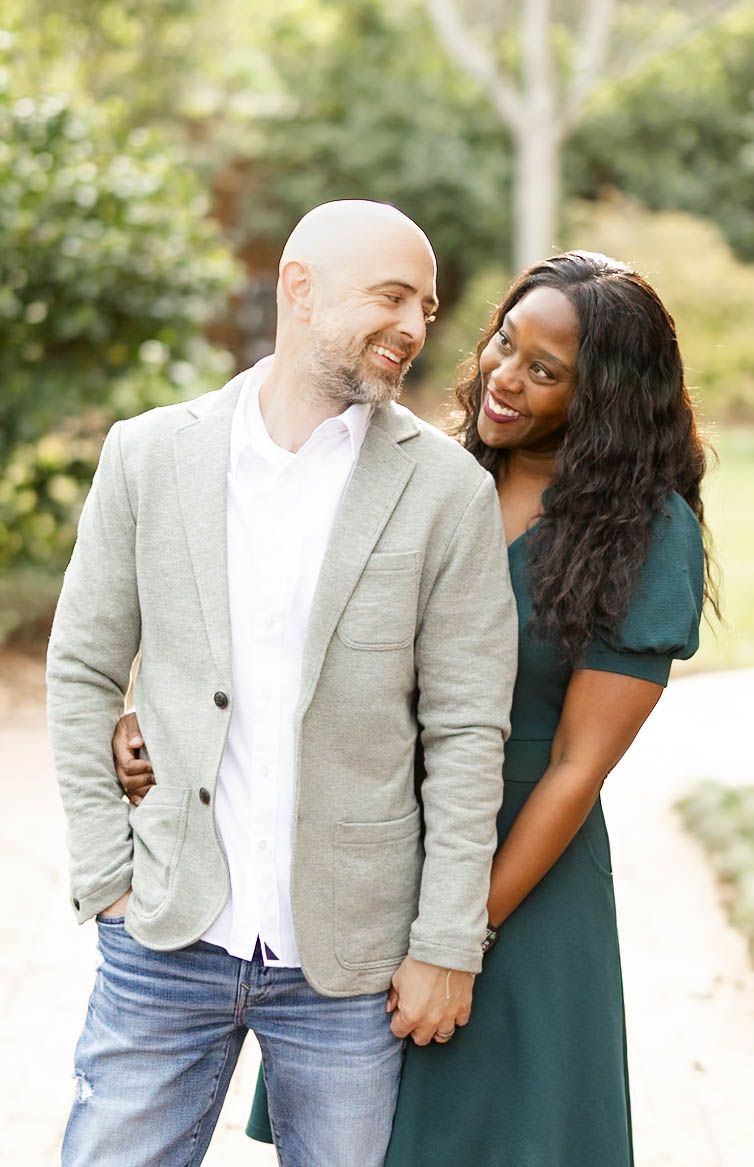 Smiling interracial couple holding hands and laughing together outdoors in a lush green park setting.