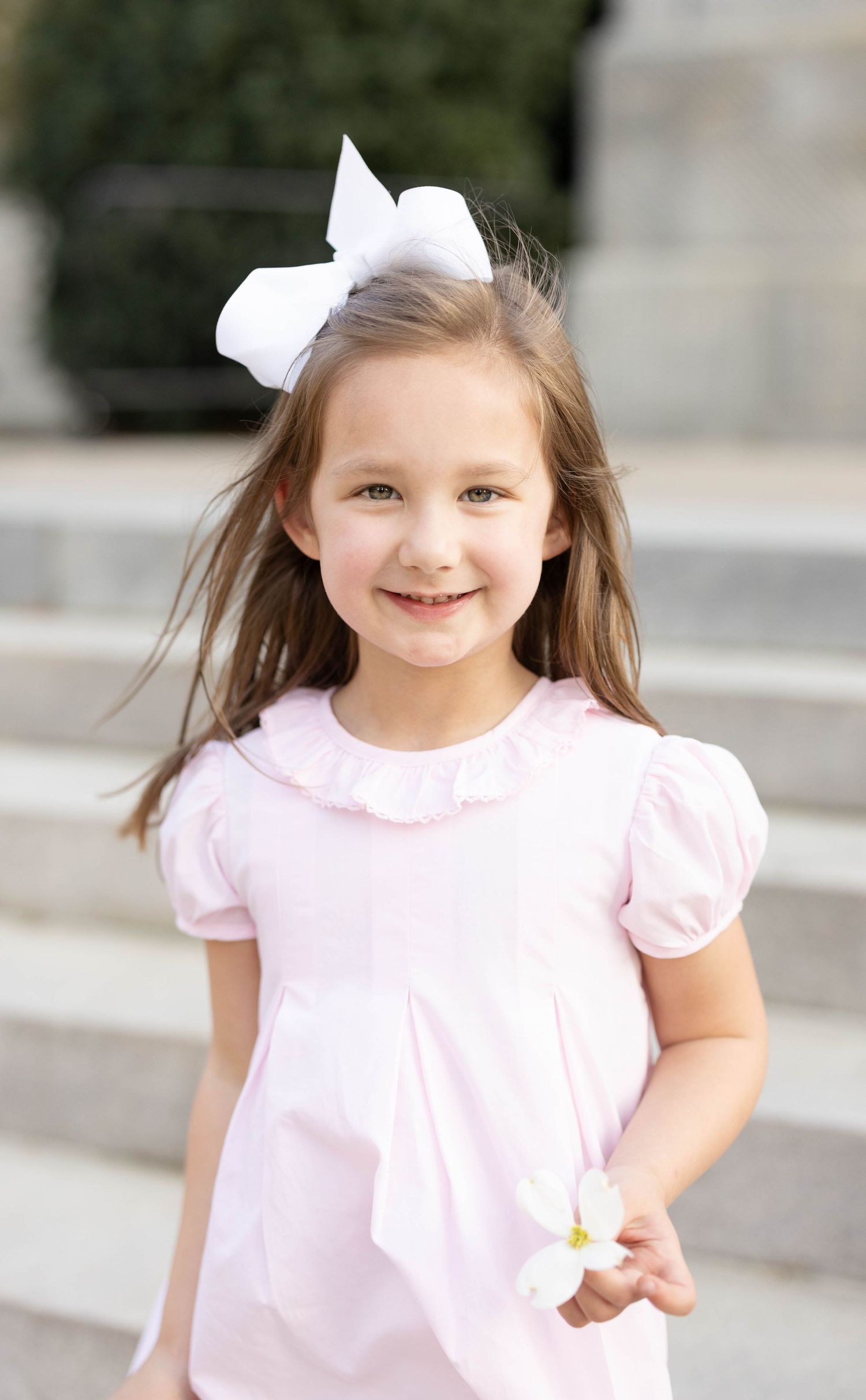 Sweet young girl in pink puff sleeve dress with large white hair bow holds a flower.