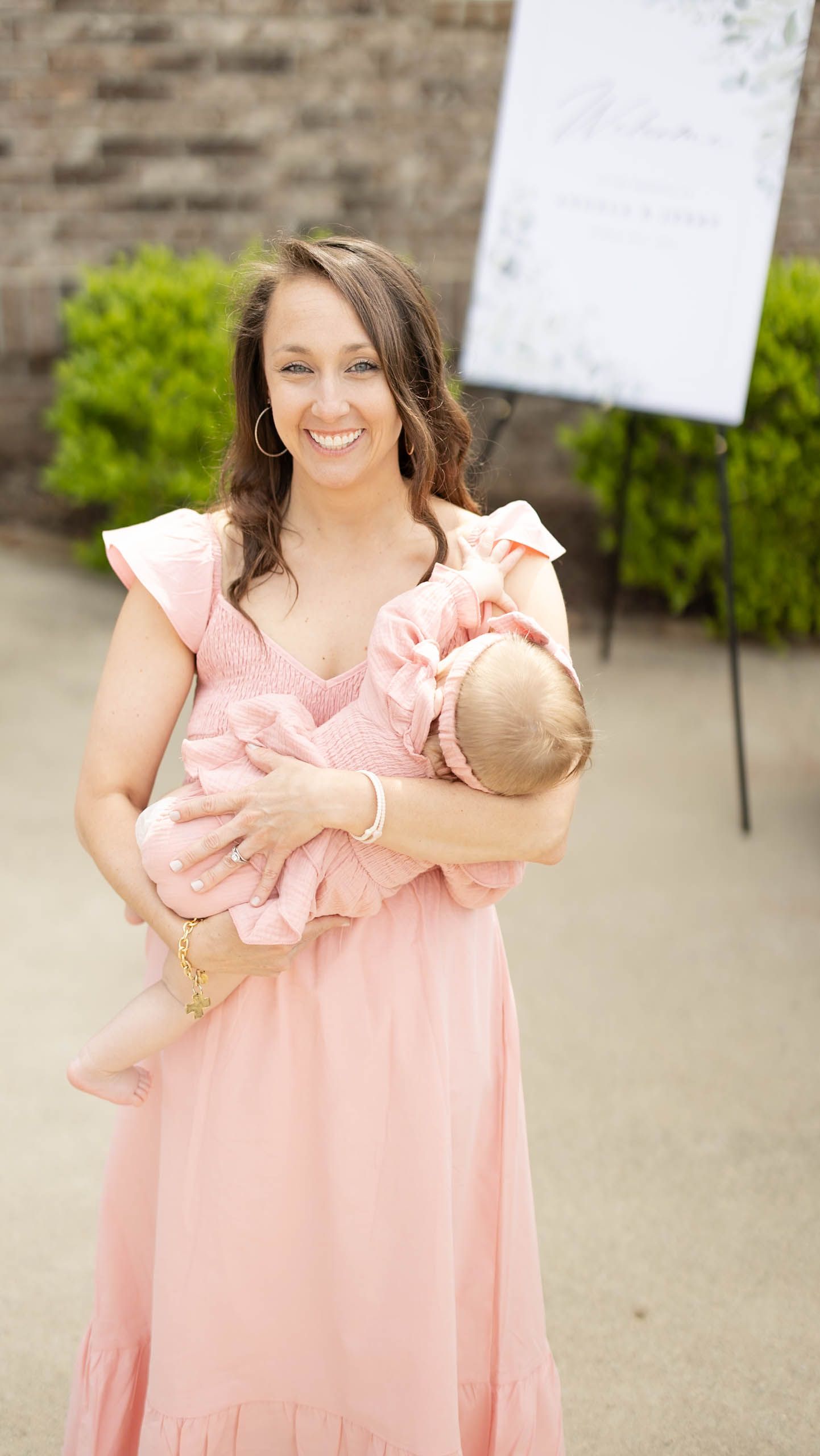A smiling woman in a pink ruffled dress holds a sleeping baby at an outdoor event near a floral sign.
