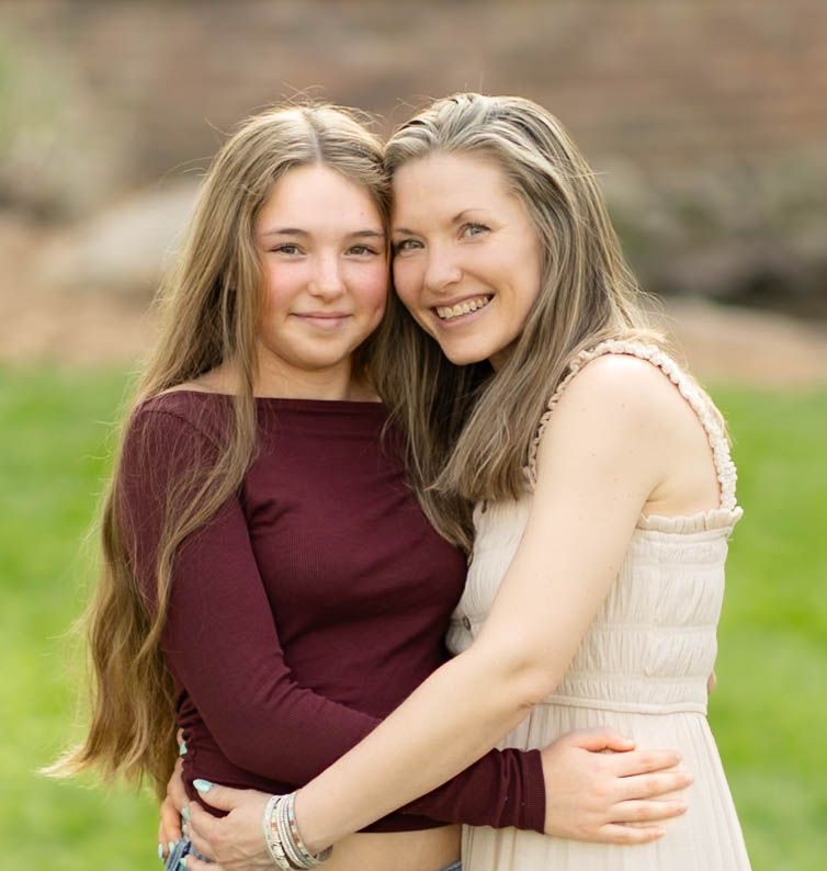 Mother and teenage daughter with long brown hair embracing and smiling together outdoors in nature.
