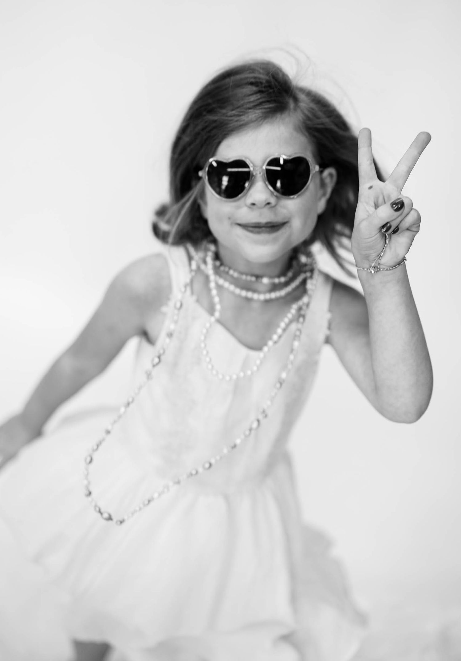 A young girl in a white dress and pearl necklace flashes a peace sign while wearing aviator sunglasses.