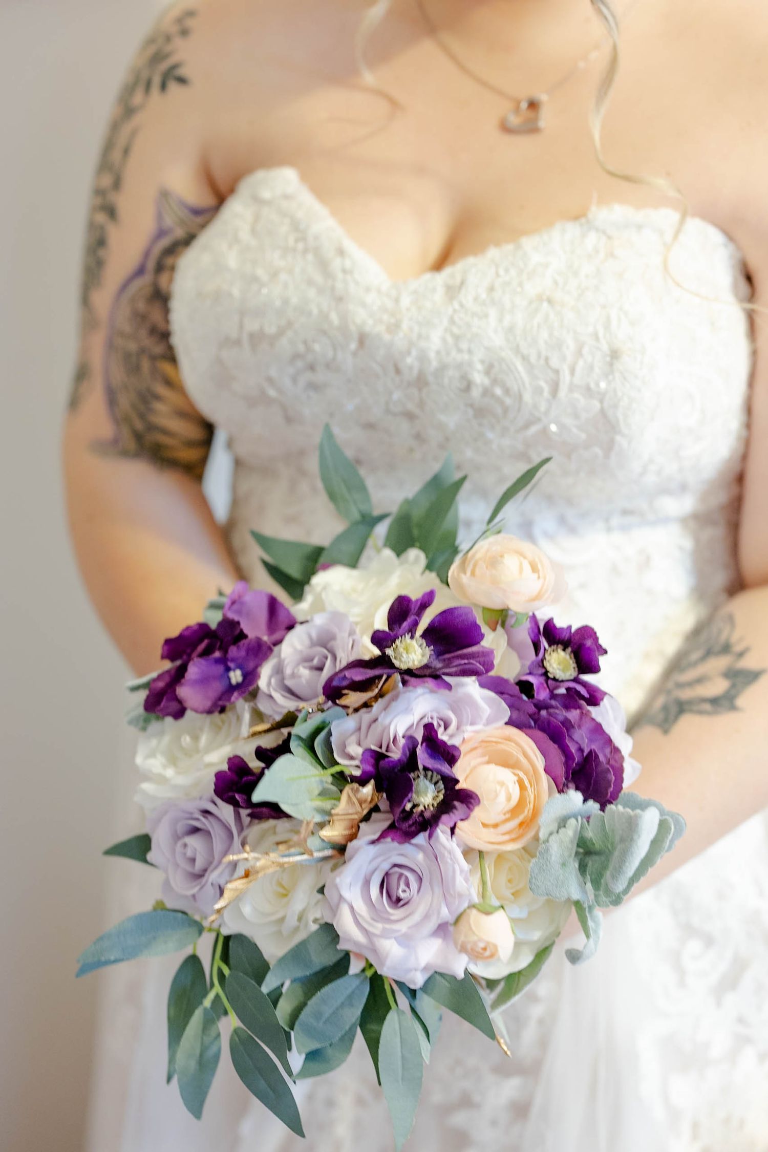 Bride in lace strapless wedding gown with tattoos holding purple, lavender, and peach bridal bouquet with greenery.