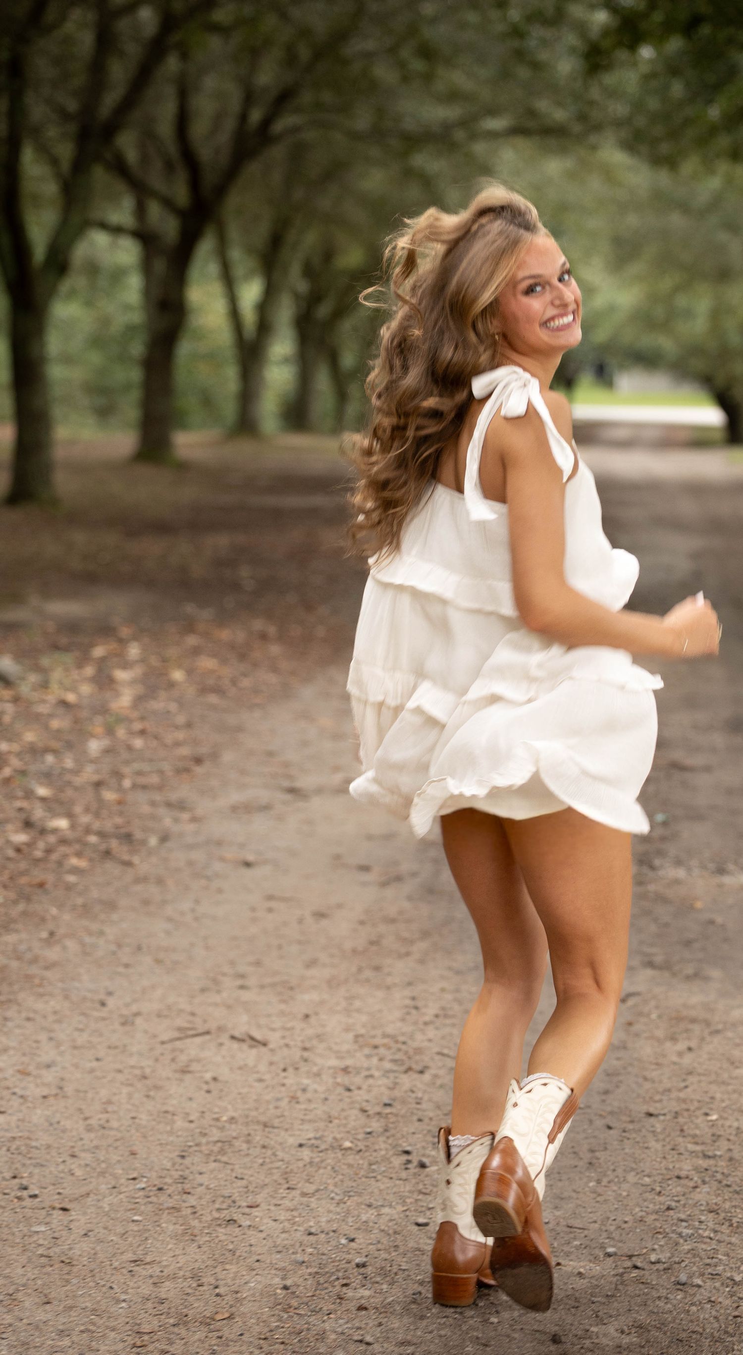Smiling woman with long wavy hair runs down a dirt path lined with trees in a white tiered dress and brown cowboy boots.