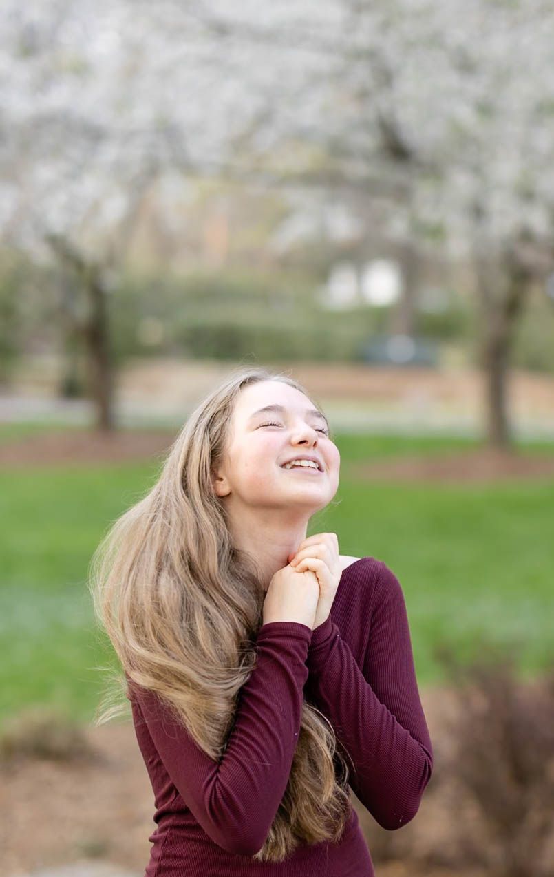 A teenage girl with long hair smiles joyfully with eyes closed under blooming spring trees.
