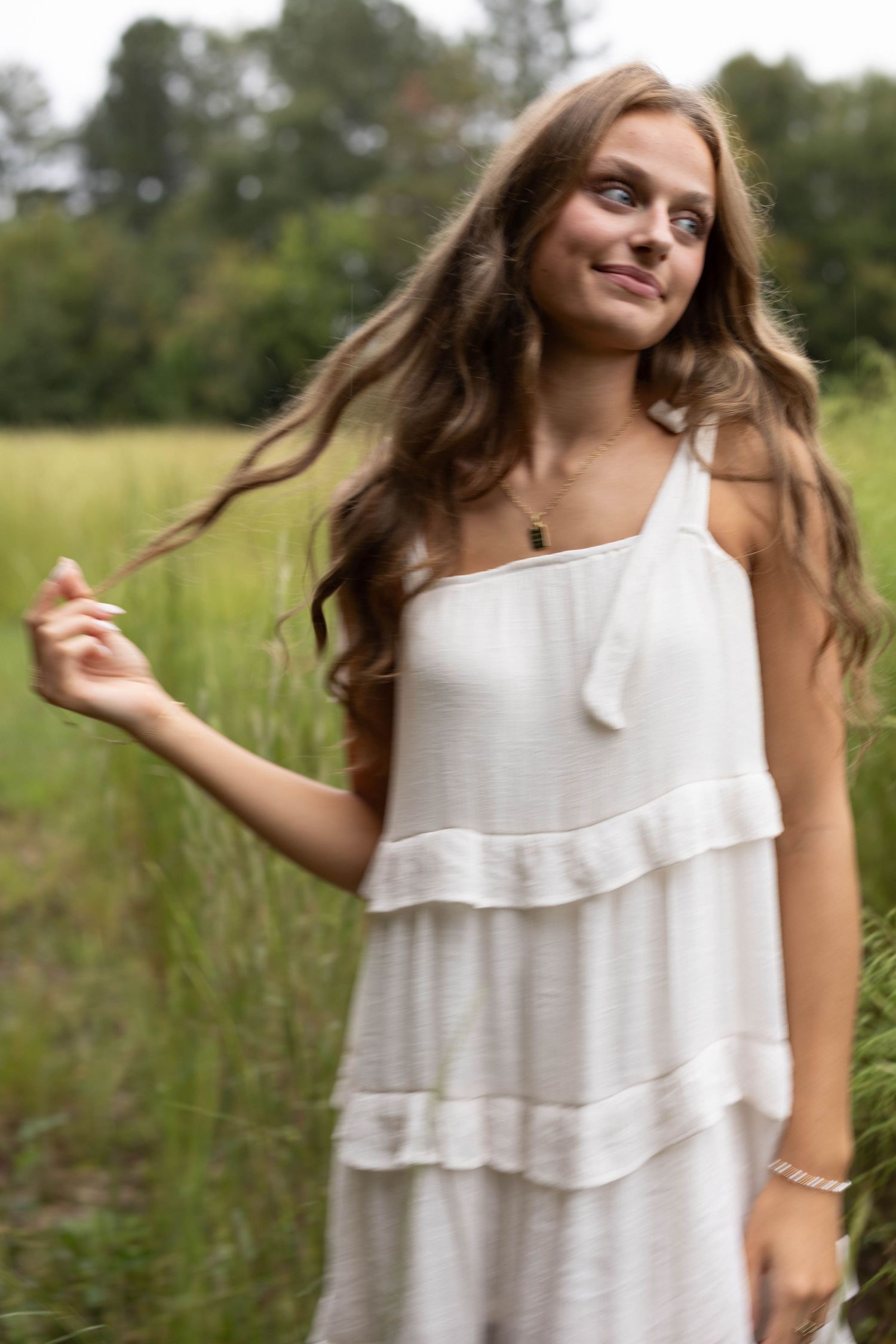 Smiling young woman with long wavy brown hair in a white tiered sundress stands in a green grassy field, gently touching her hair.
