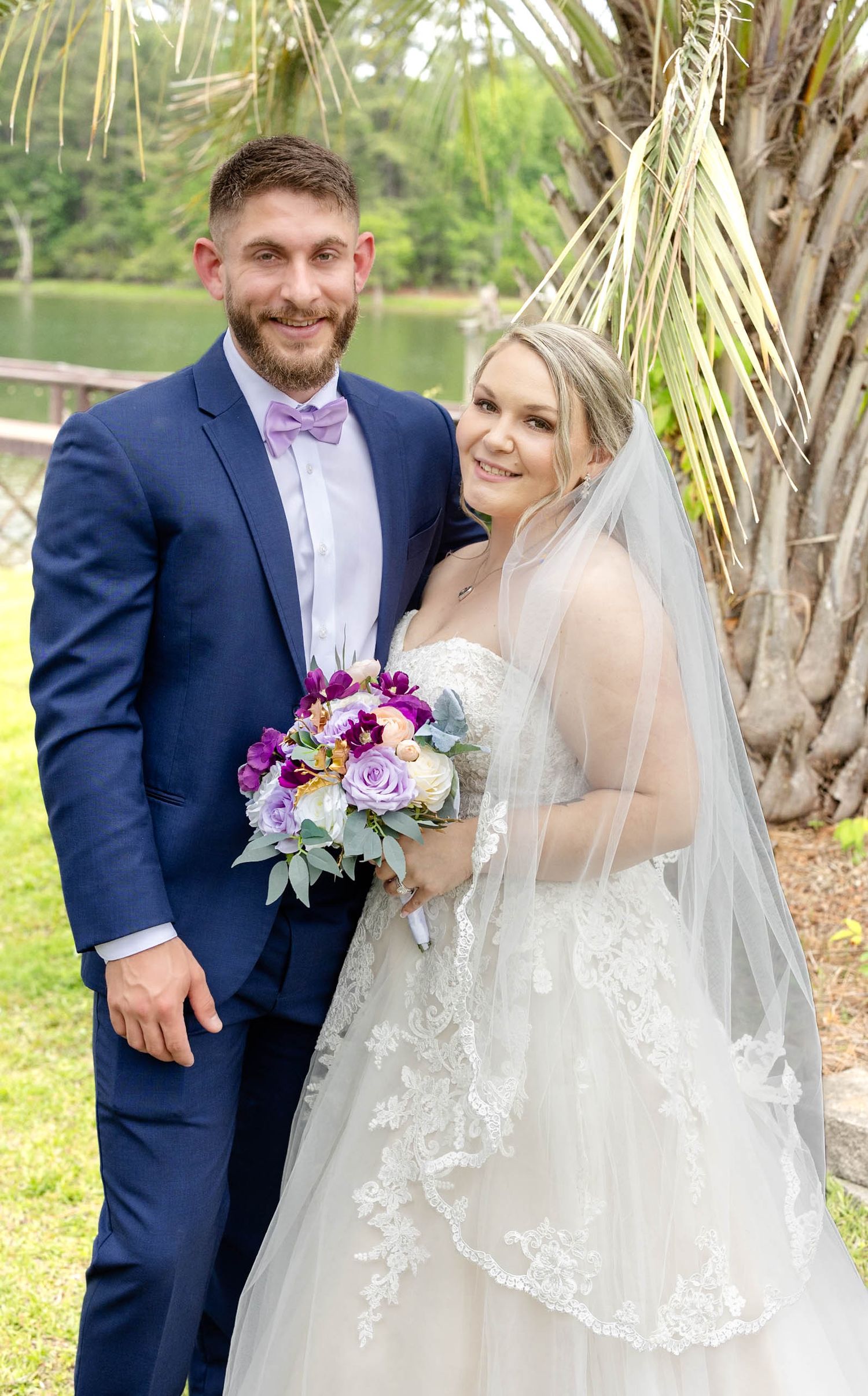 Bride in lace gown with veil holding purple bouquet stands beside groom in navy suit with lavender bow tie outdoors.
