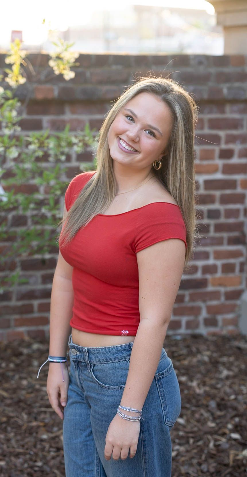 Smiling teenage girl in red off-shoulder crop top and jeans posing in front of a brick wall.