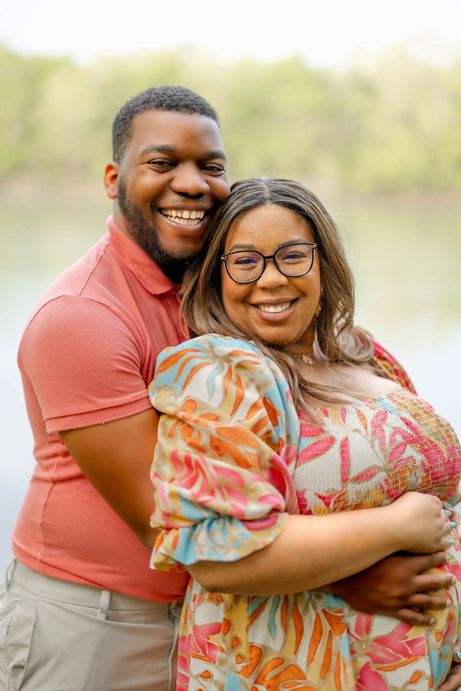 Joyful expecting couple embracing and smiling together outdoors near a serene lake backdrop.