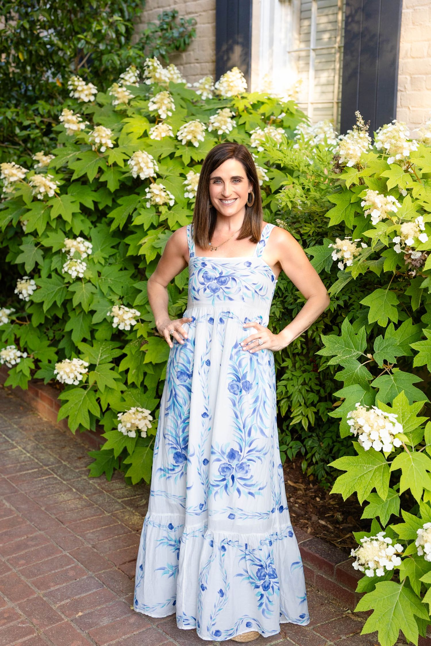 Woman in blue floral maxi dress posing in front of lush green bushes with white blooms on brick path.