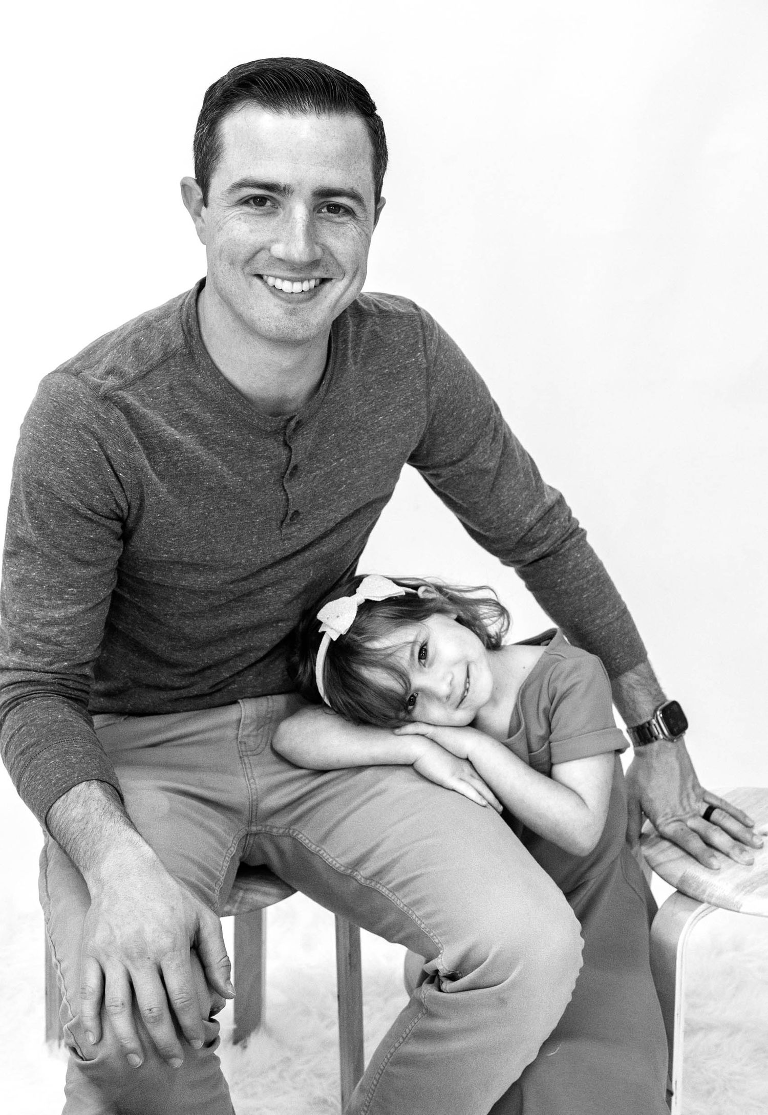 Black and white portrait of a smiling man sitting on a stool with a young girl resting her head on his lap.