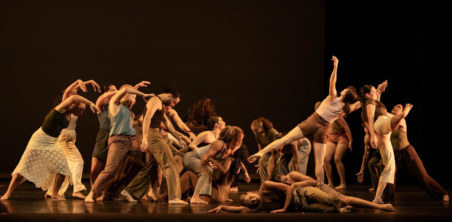 A group of contemporary dancers in casual clothing perform an expressive choreographed piece on a dimly lit stage.