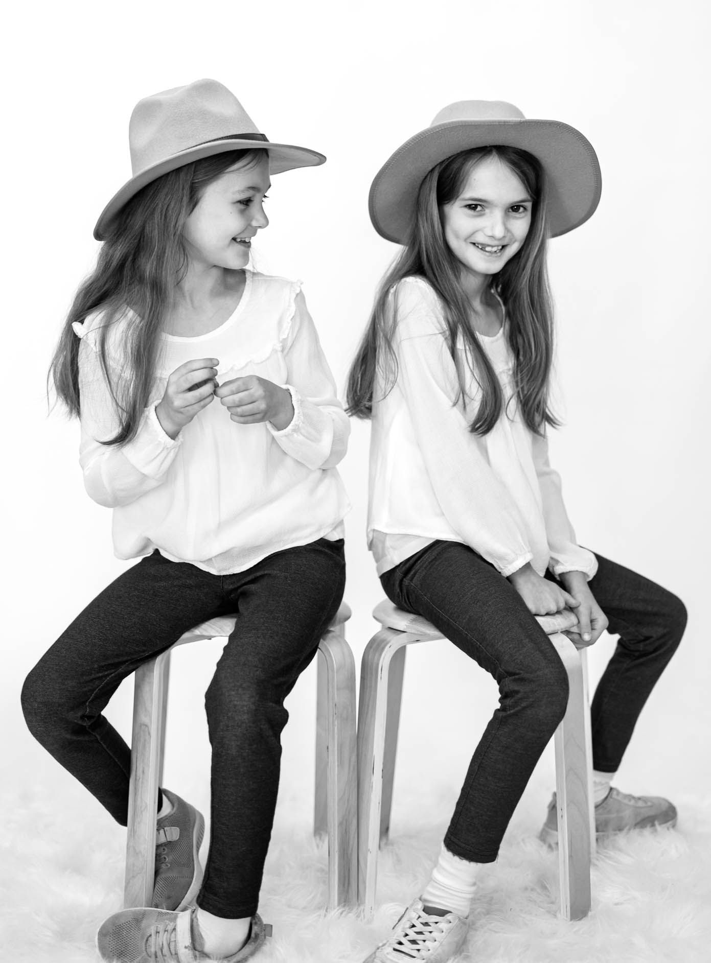 Two young girls in matching white tops, dark jeans, and wide-brim hats sit on wooden stools in a black and white photo.