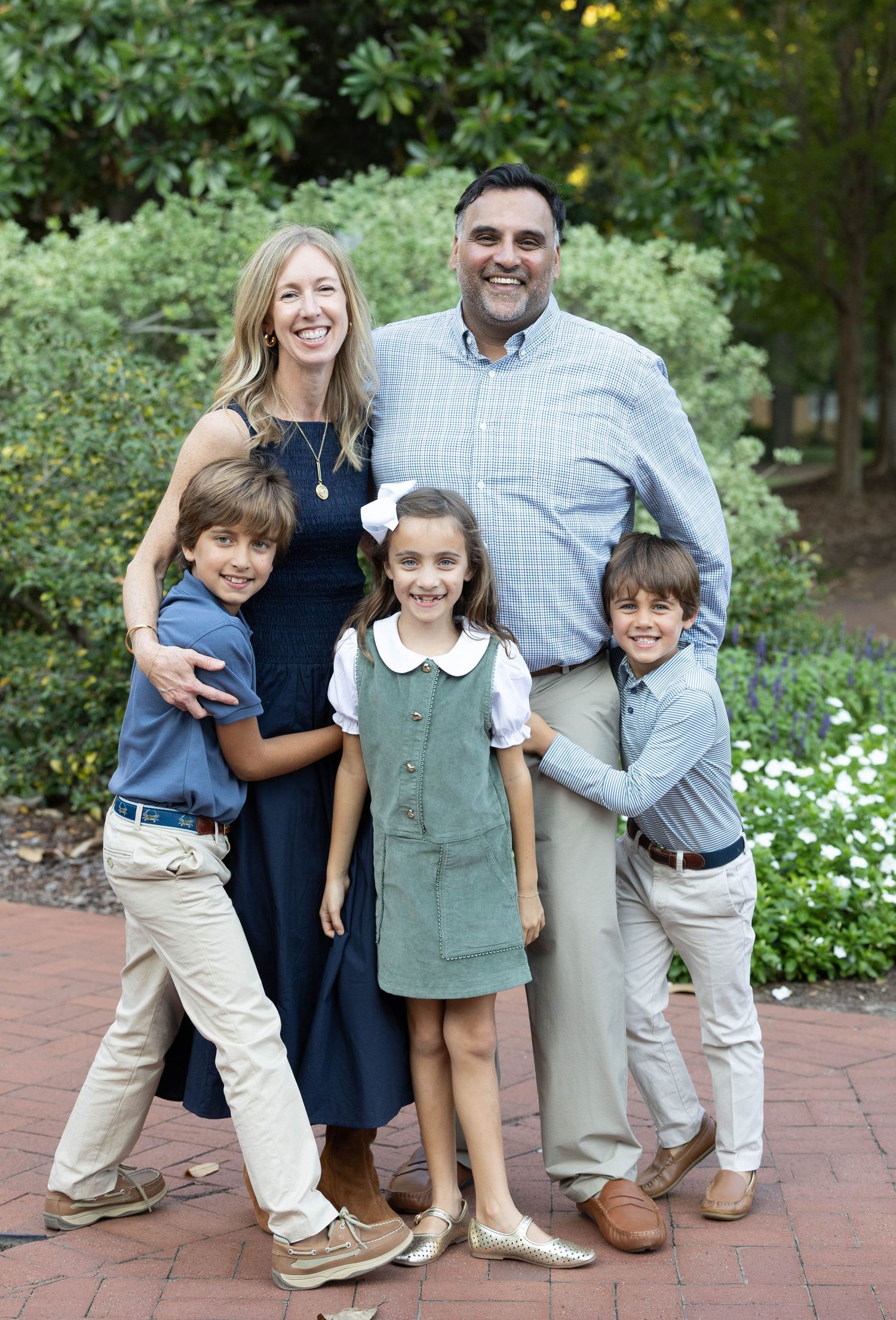 A smiling family of five poses outdoors on a brick patio, with two parents and three children in front of lush green shrubs.