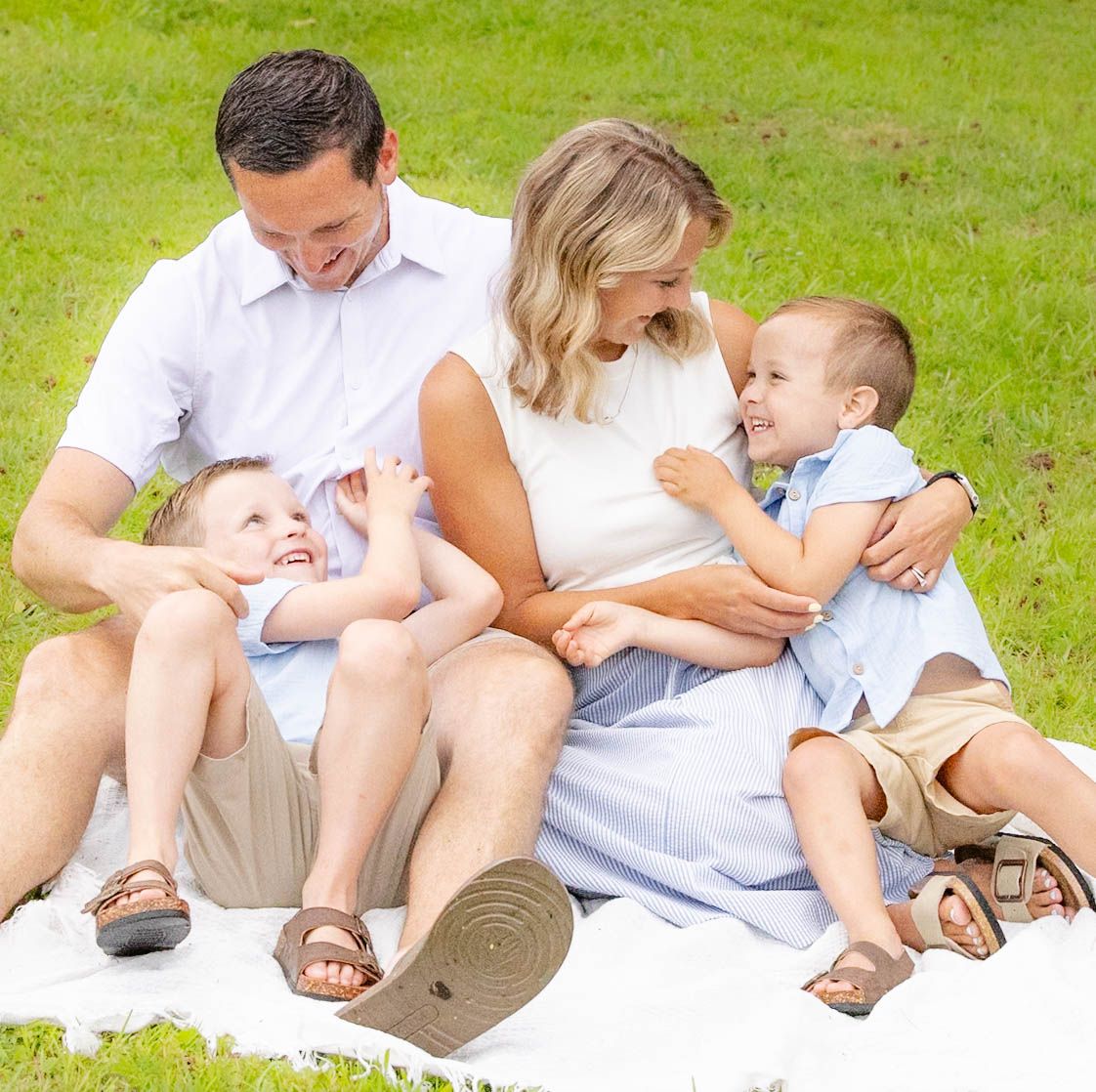 Happy family of four sitting on a white blanket in a green park, parents smiling with two young children.